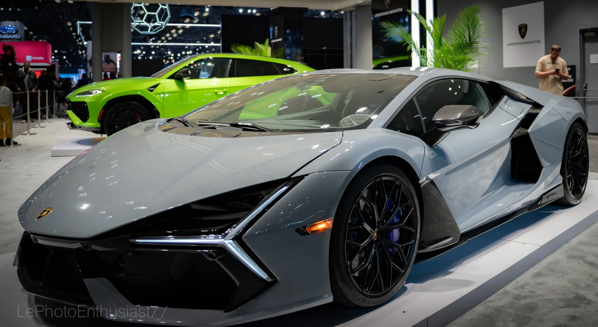 A silver Lamborghini sports car and a green Lamborghini sports car on display at an auto show, with people in the background.