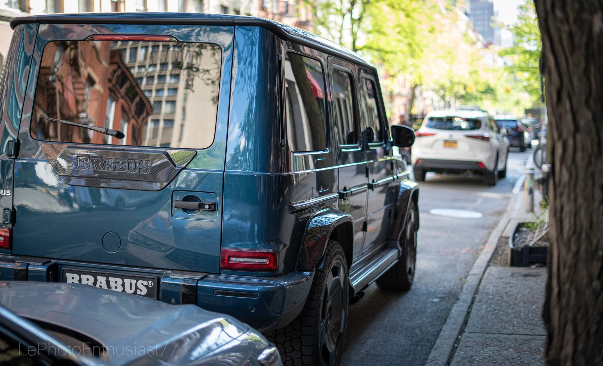 A blue Brabus modified luxury SUV parked on a city street, with other cars and trees in the background.