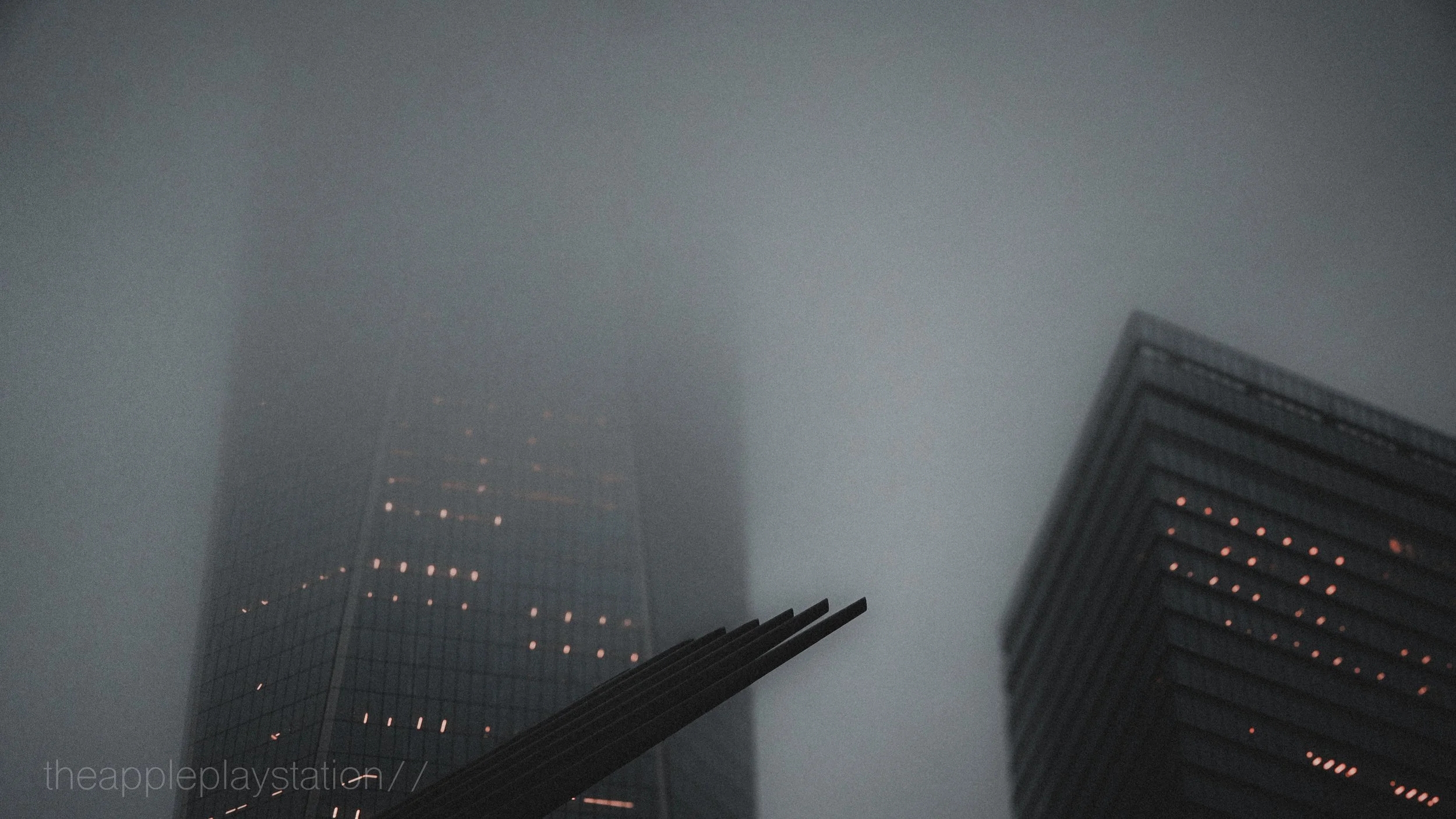 Faint view of two tall buildings with grid-like windows, shrouded in fog, one taller and in the background, the other shorter on the right with illuminated windows.