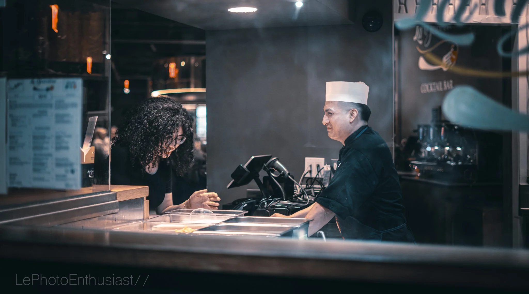 A woman with curly hair and glasses is paying at a restaurant counter, while a chef in a white hat and black uniform stands behind the counter smiling.