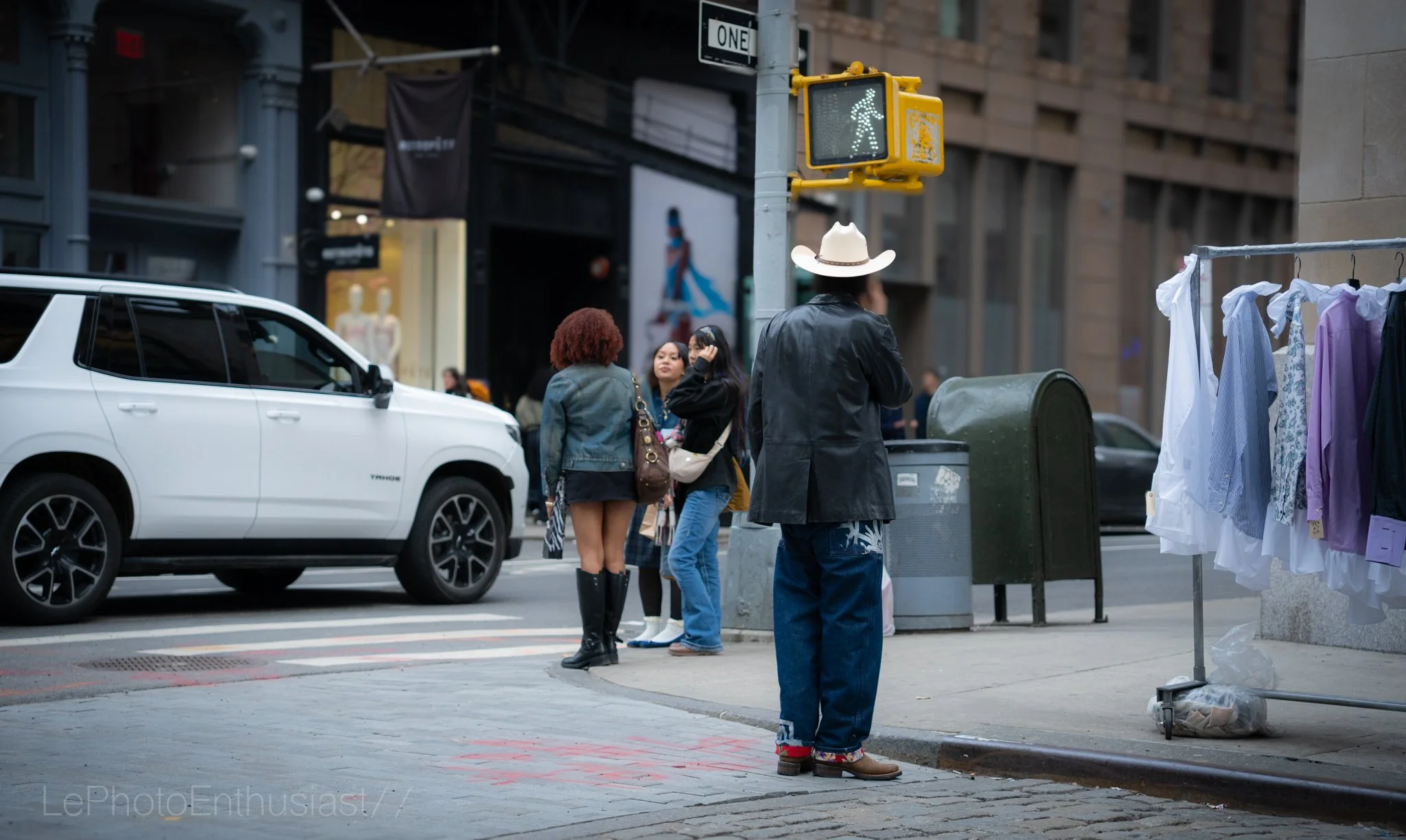 People crossing a city street at a crosswalk with a pedestrian signal showing a walking figure, a white SUV parked nearby, a clothing rack with hanging garments, and a street sign that says "ONE".