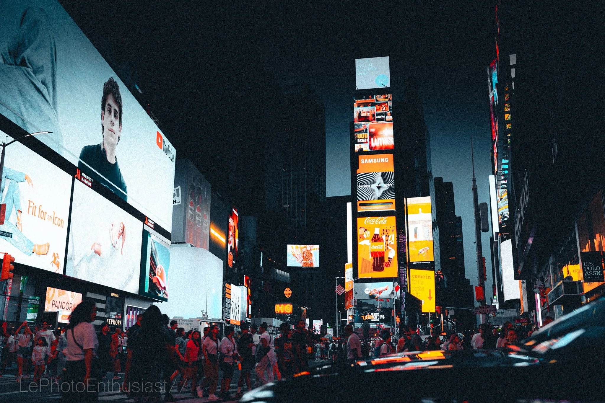 Bright digital billboards and advertisements illuminating Times Square in New York City at night, with a crowd of people walking across the street.