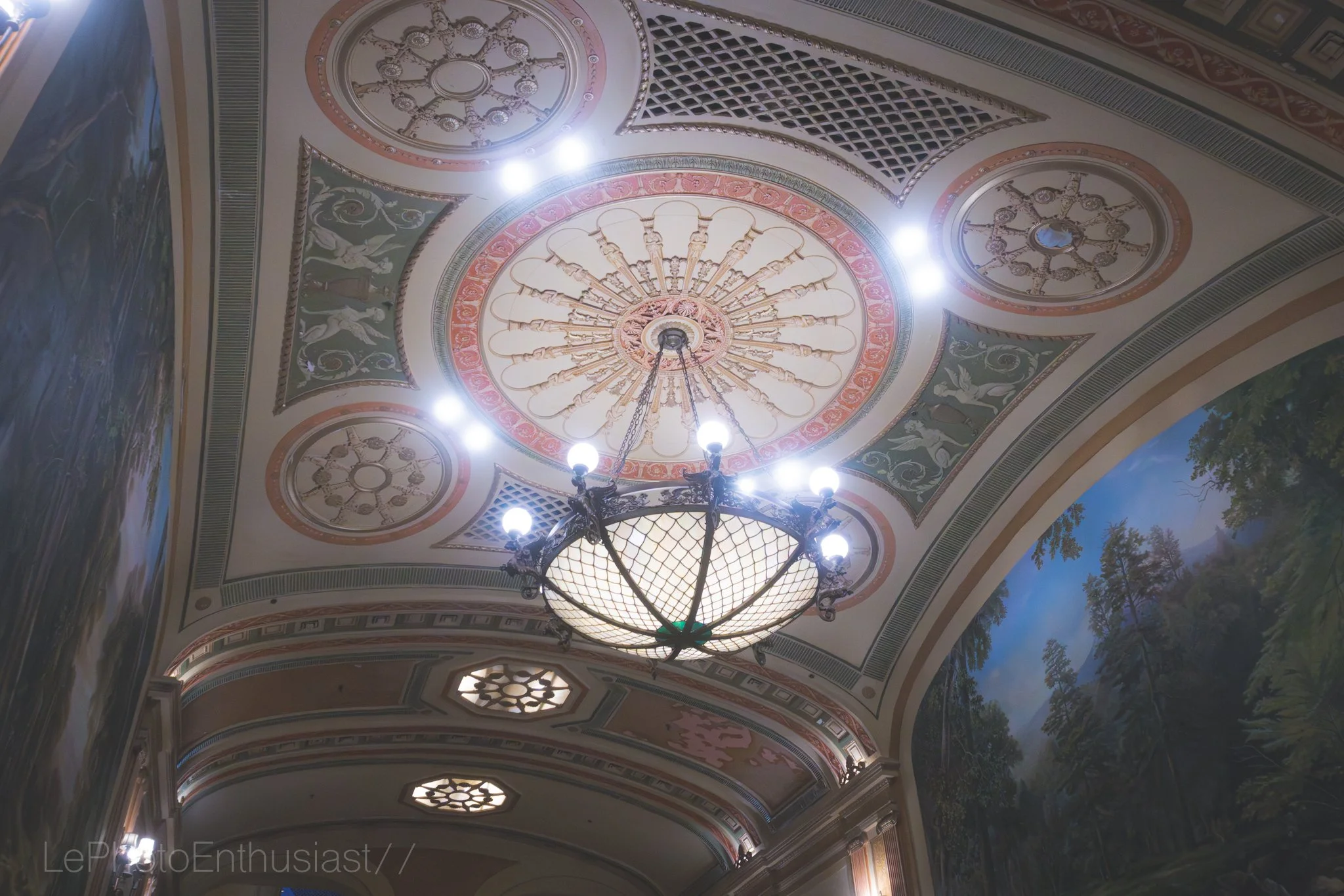 Ornate ceiling with decorative patterns, a chandelier with multiple lights, and painted murals of landscape scenes.