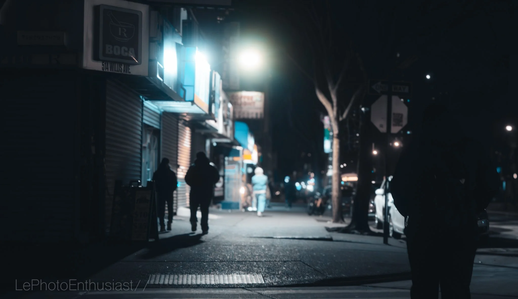 Nighttime city street scene with illuminated storefronts, a few pedestrians walking along the sidewalk, parked cars, and streetlights.