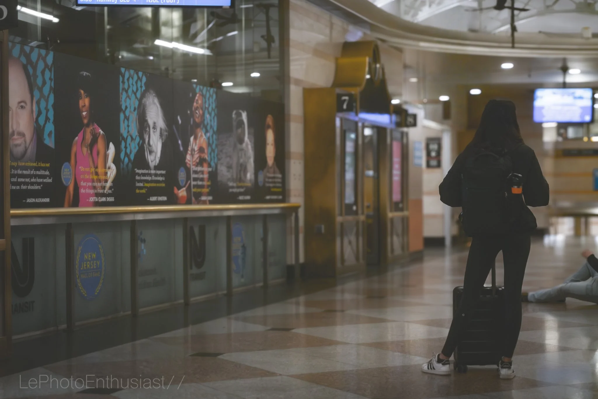 A woman with a black backpack and rolling suitcase stands in an airport terminal, looking at a row of posters featuring portraits of notable people.