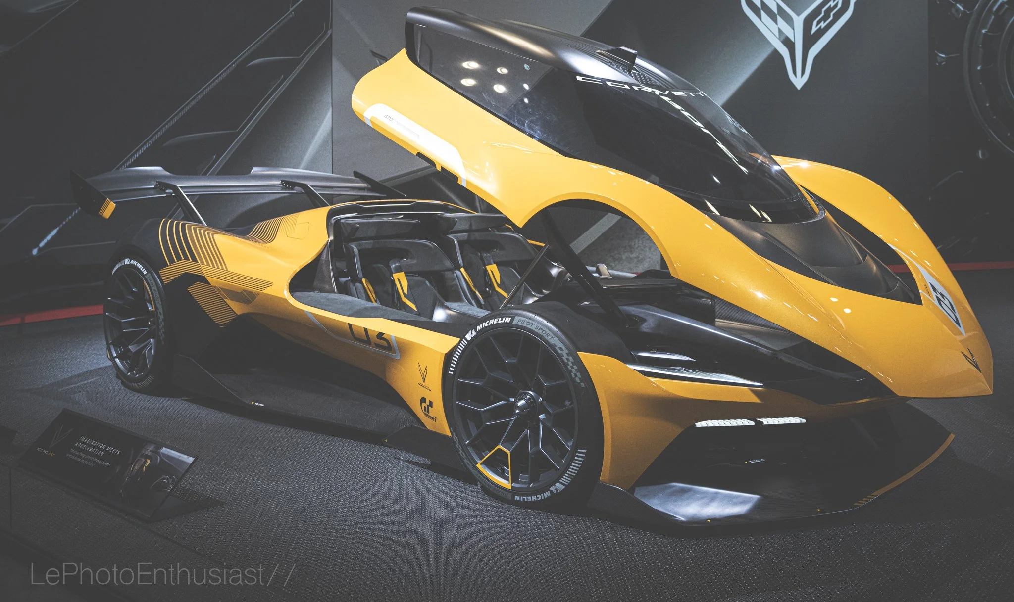 A yellow and black sports car with racing decals on display at an auto show. The car has a sleek, aerodynamic design and is surrounded by a dark environment with a display plaque at its front.