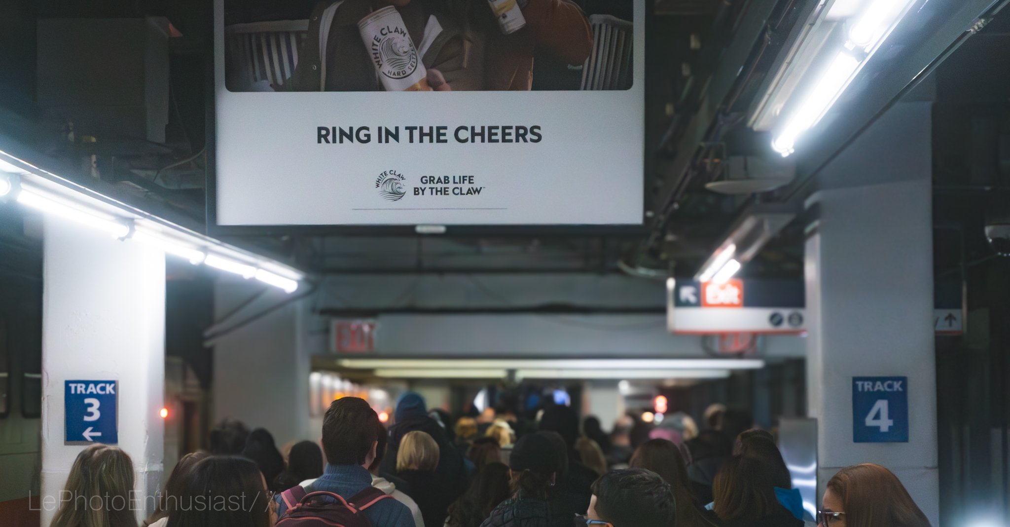 Crowded subway station with people waiting on platform under signs indicating tracks 3 and 4, illuminated overhead lighting, and a sign promoting White Claw with the slogan 'Ring in the Cheers.'