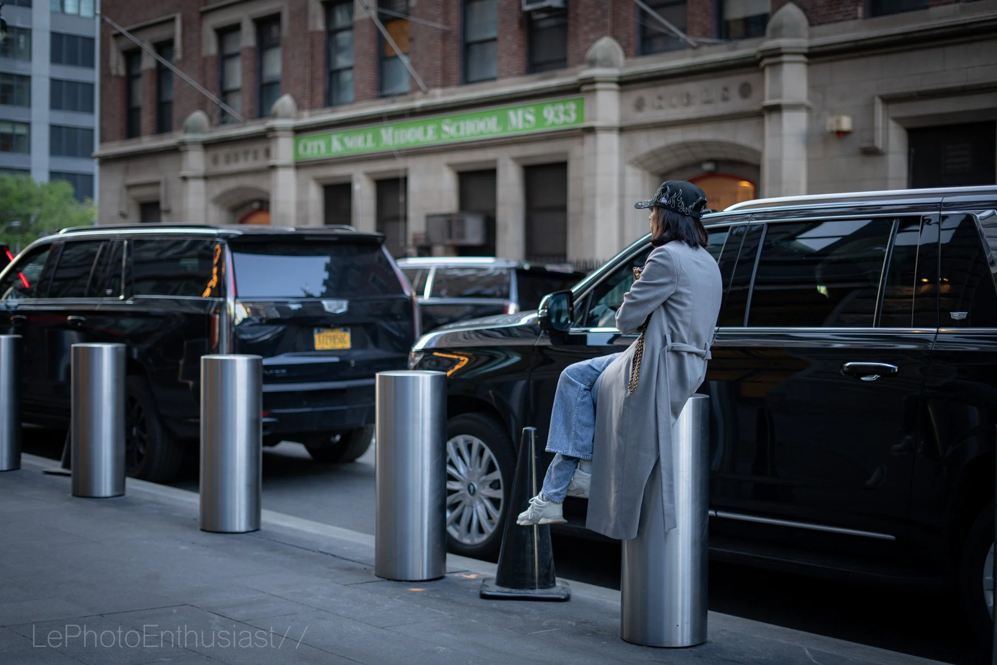 A woman wearing a black cap and gray coat sitting on a black car parked on the street, with a background of an old brick building with a green sign reading 'CITY K VIRL MIDDLE SCHOOL NIS 953' and other parked cars.