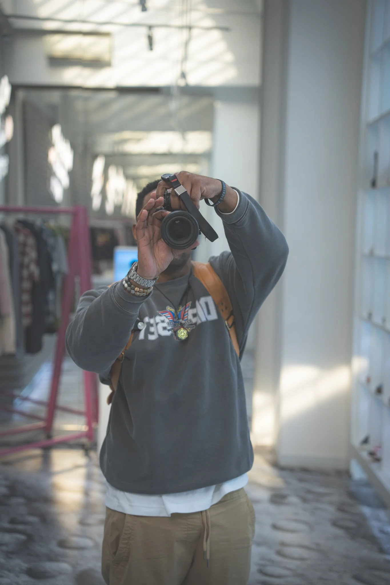 Person taking a photo with a camera in a bright indoor space, wearing a dark hoodie and tan pants.