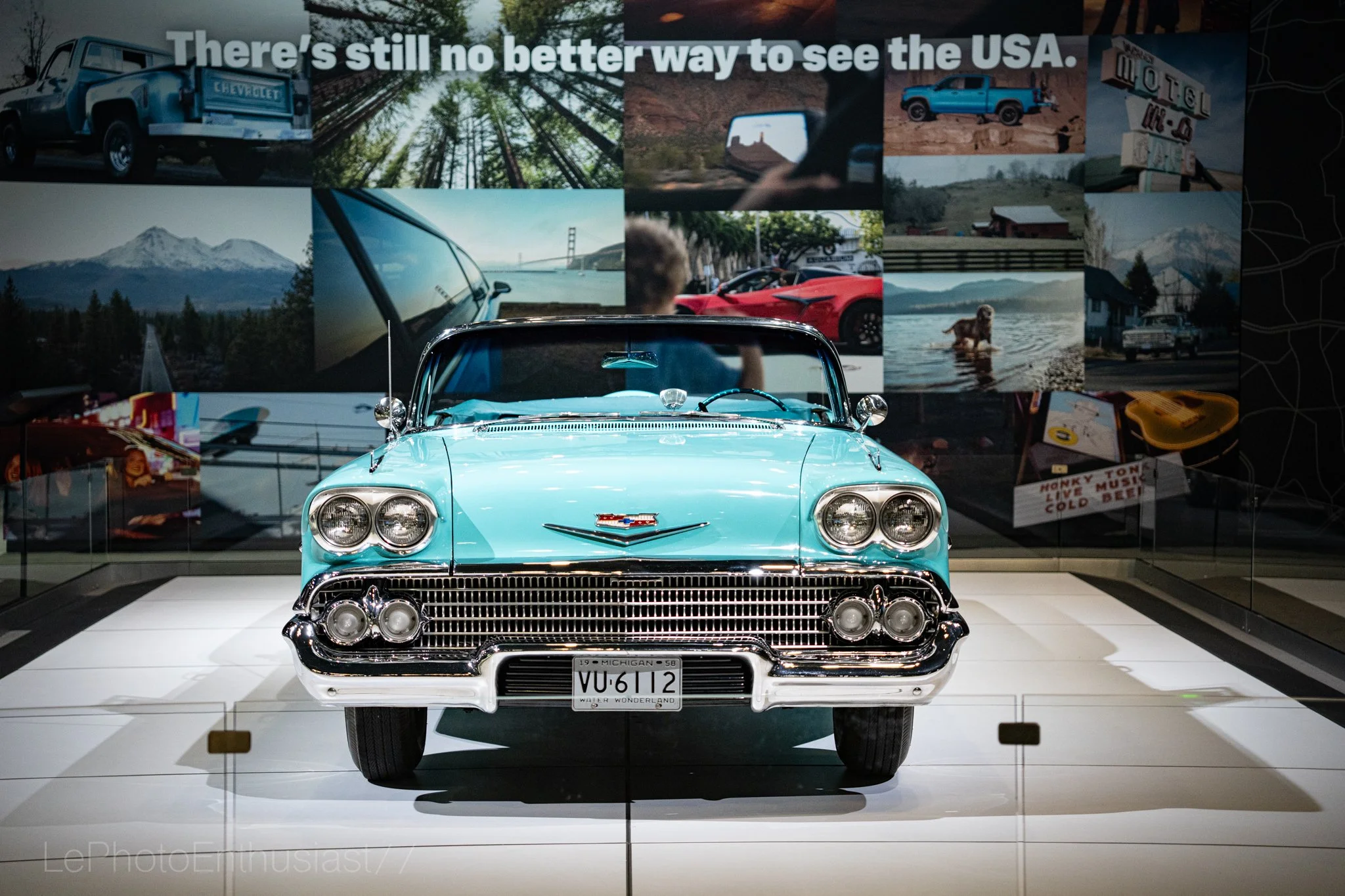 A classic light blue 1958 Chevrolet car displayed in a museum with a collage of images in the background, including mountains, a bridge, nature scenes, vehicles, a dog, and signs, along with the text 'There's still no better way to see the USA.'
