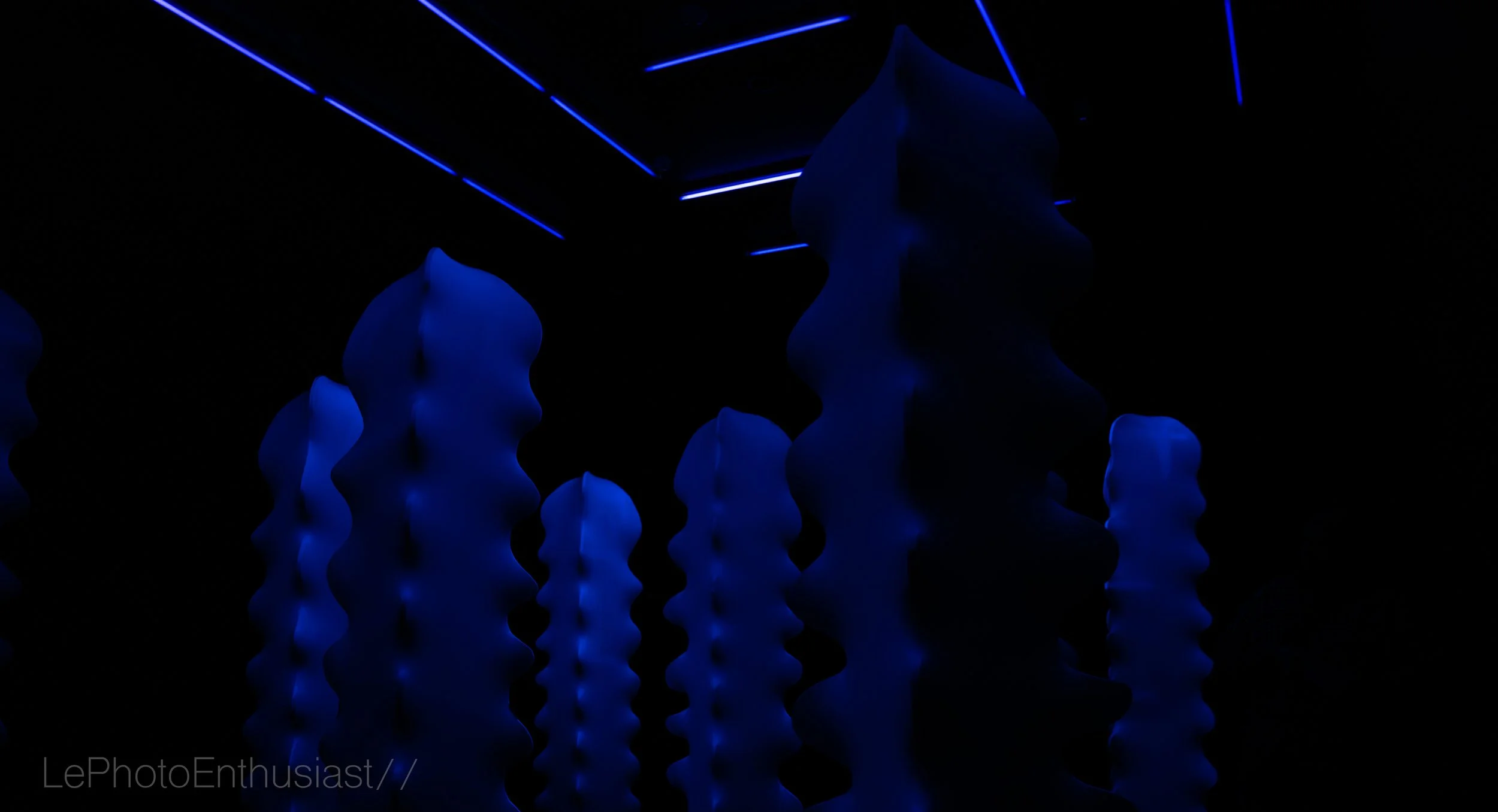 Interior of a dark room with blue LED strip lights on the ceiling and illuminated abstract sculptures or objects with wavy shapes.