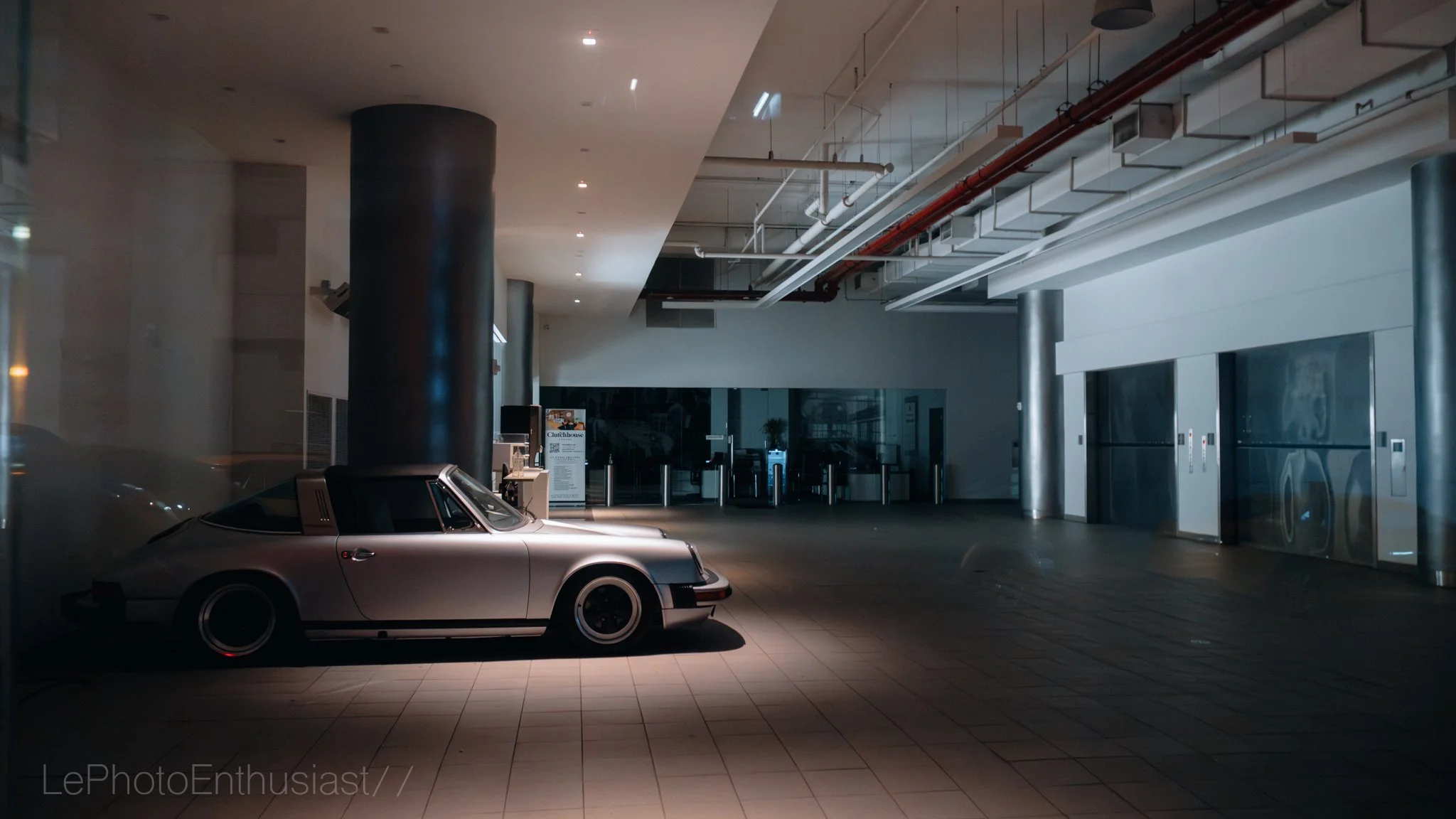 A vintage silver Porsche car parked inside a modern, dimly-lit building with elevator doors on the right and industrial piping on the ceiling.