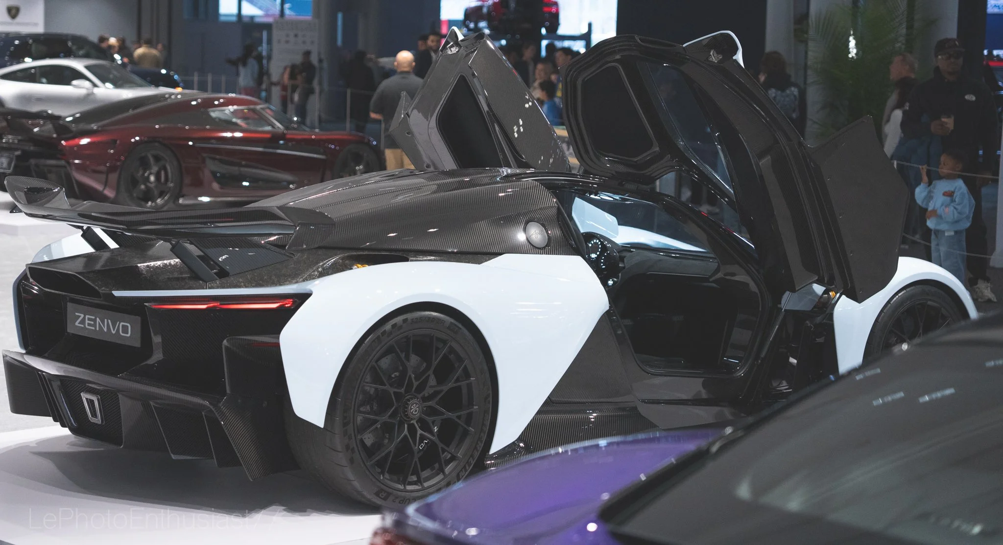 A white and black sports car with scissor doors open at an auto show, with other cars and people in the background.