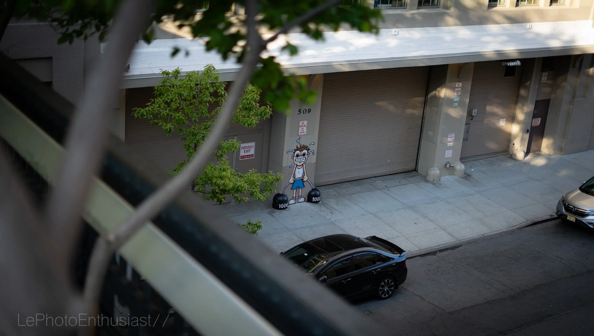 A cartoon street art of a muscular man lifting a barbell with dumbbells labeled 1000 lb. in front of a building with the number 509, partially obscured by tree leaves and parked cars.