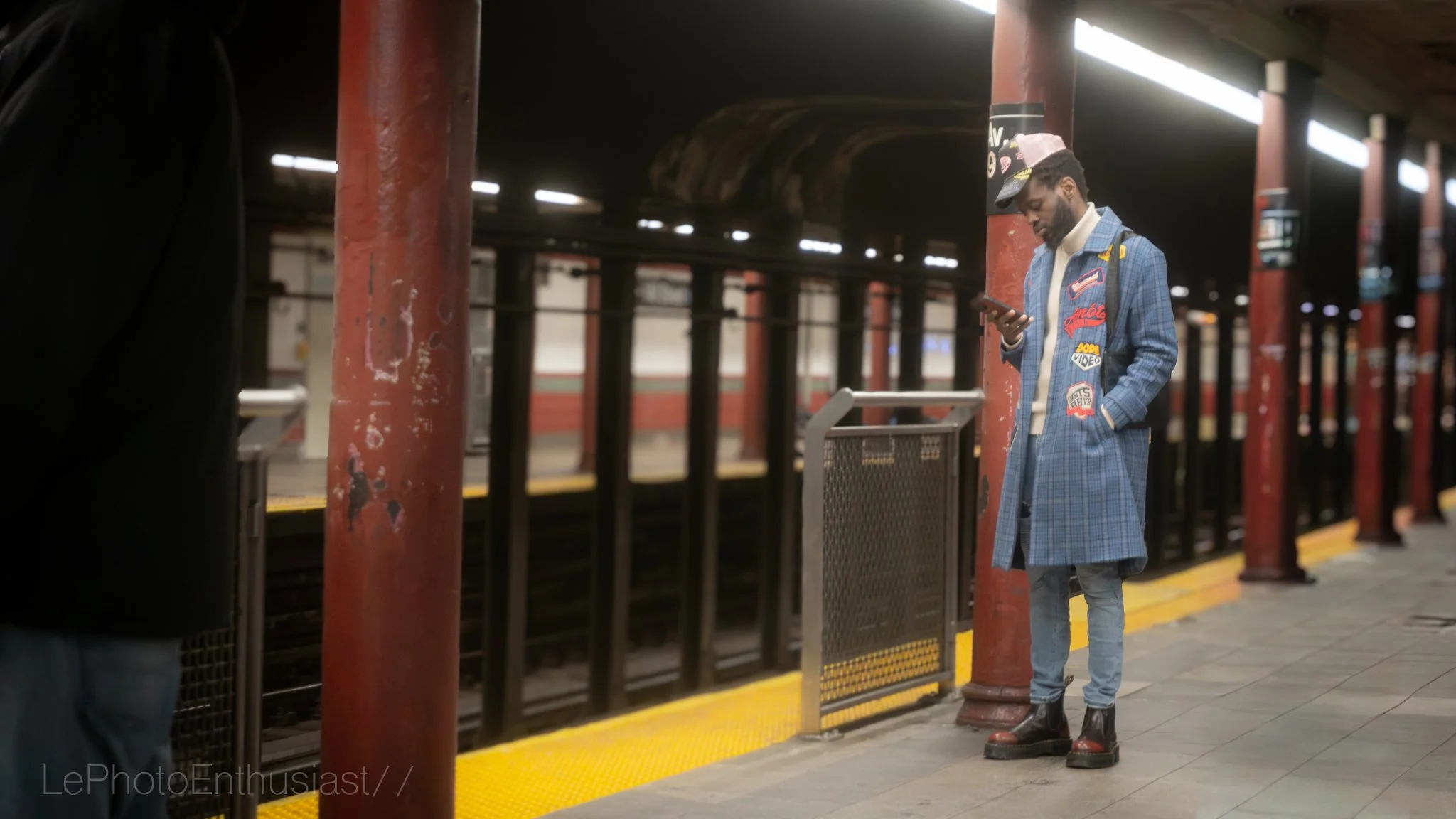 A man standing at a subway station platform, wearing a blue plaid coat with patches, a cap, and black boots, looking at his phone. The station has red pillars, a yellow safety strip on the platform edge, and a dark background with some lights.