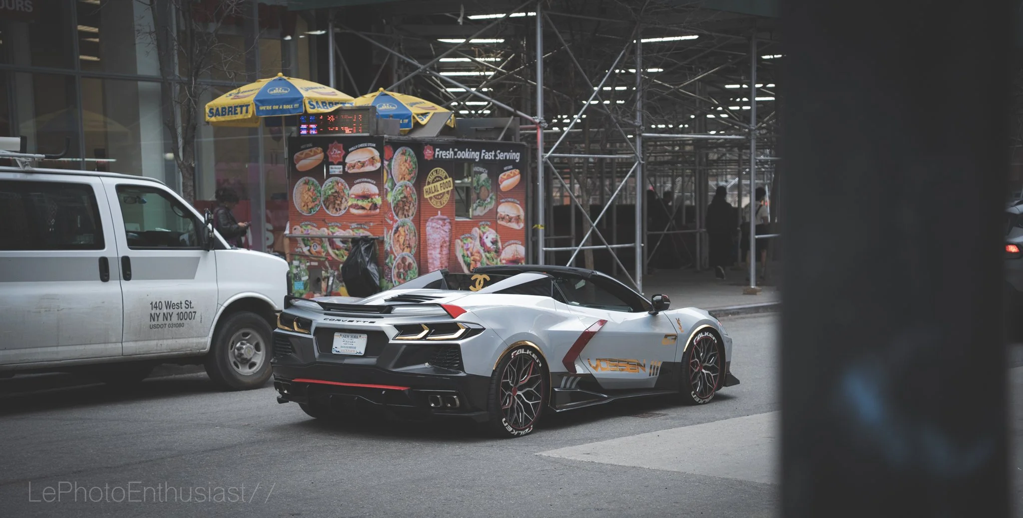 A silver sports car with Lamborghini branding parked on the street next to a white van, with a fast-food restaurant in the background.
