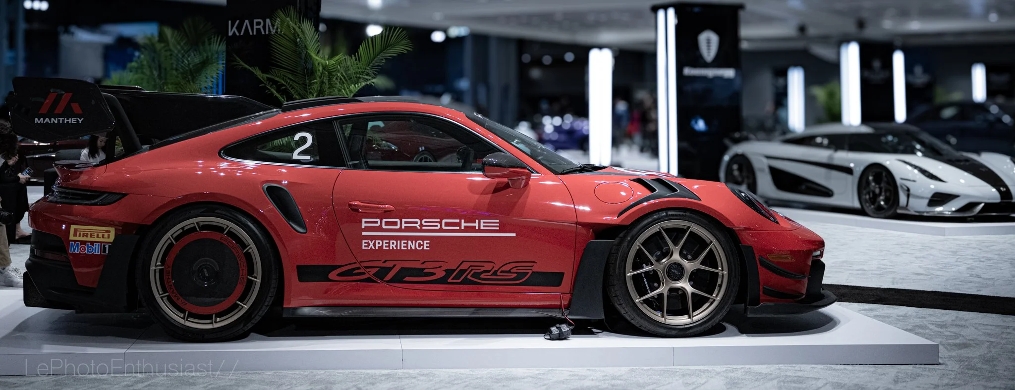 Red Porsche race car with 'Porsche Experience' and 'GT3 RS' decals displayed in an indoor showroom, surrounded by other sports cars in the background.