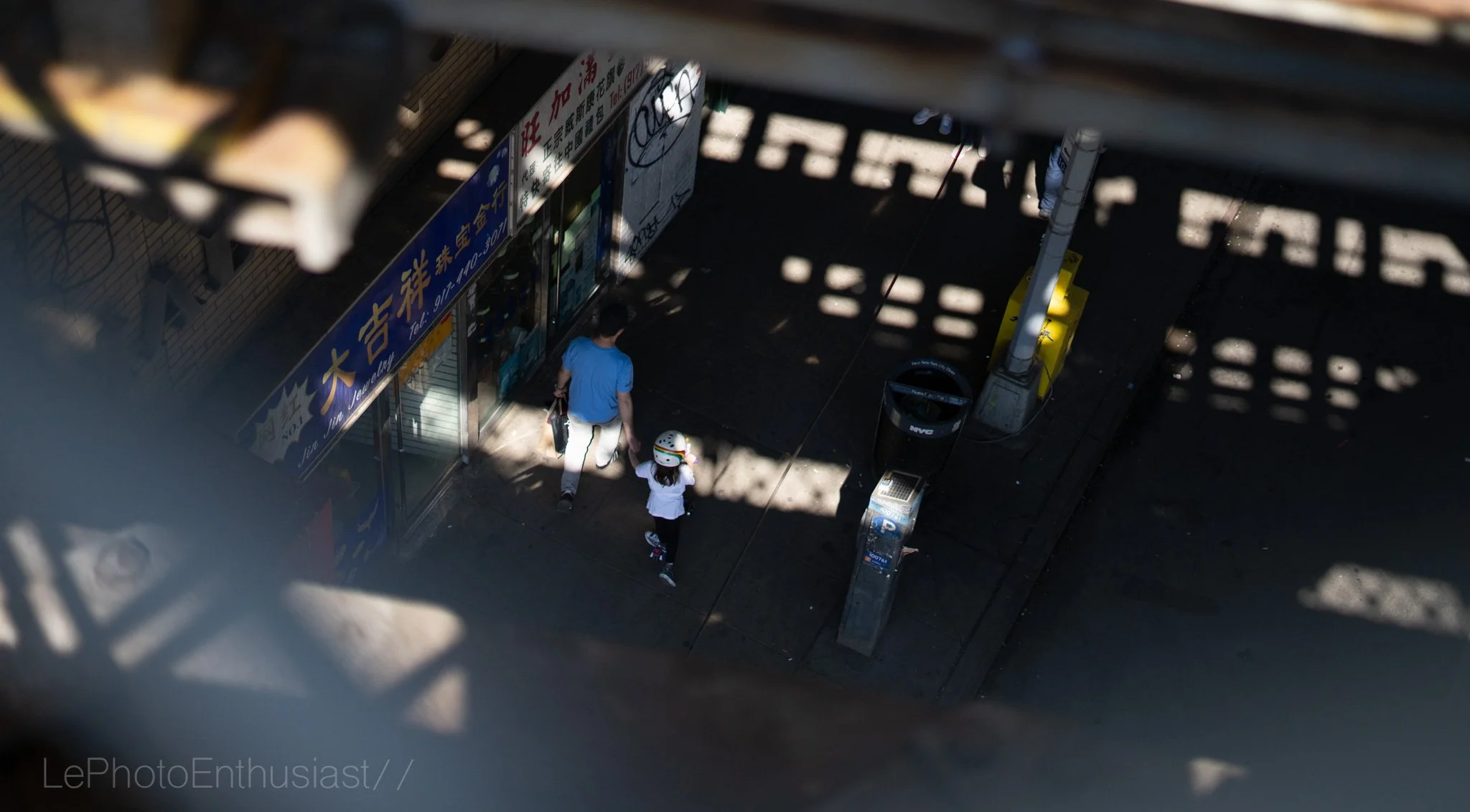 View from above of a street scene through a metal structure, showing a man and a child walking on the sidewalk near storefronts, with shadows and sunlight creating patterns on the ground.