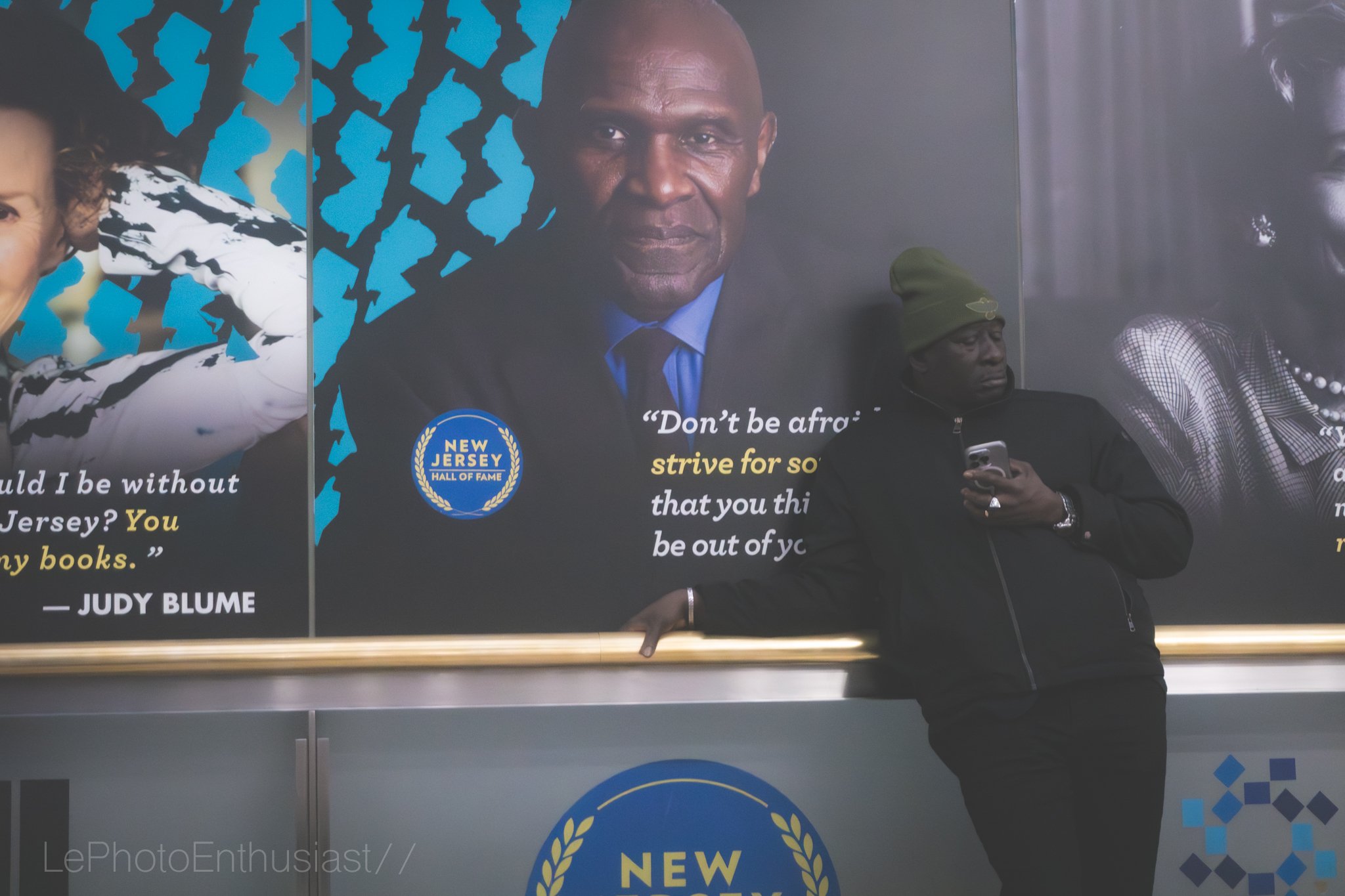 A man wearing a green beanie and black jacket is standing, leaning on a ledge, looking at his phone. Behind him is a large poster of a man with a serious expression, dressed in a suit, with a quote, a New Jersey Hall of Fame logo, and part of another poster with a woman.