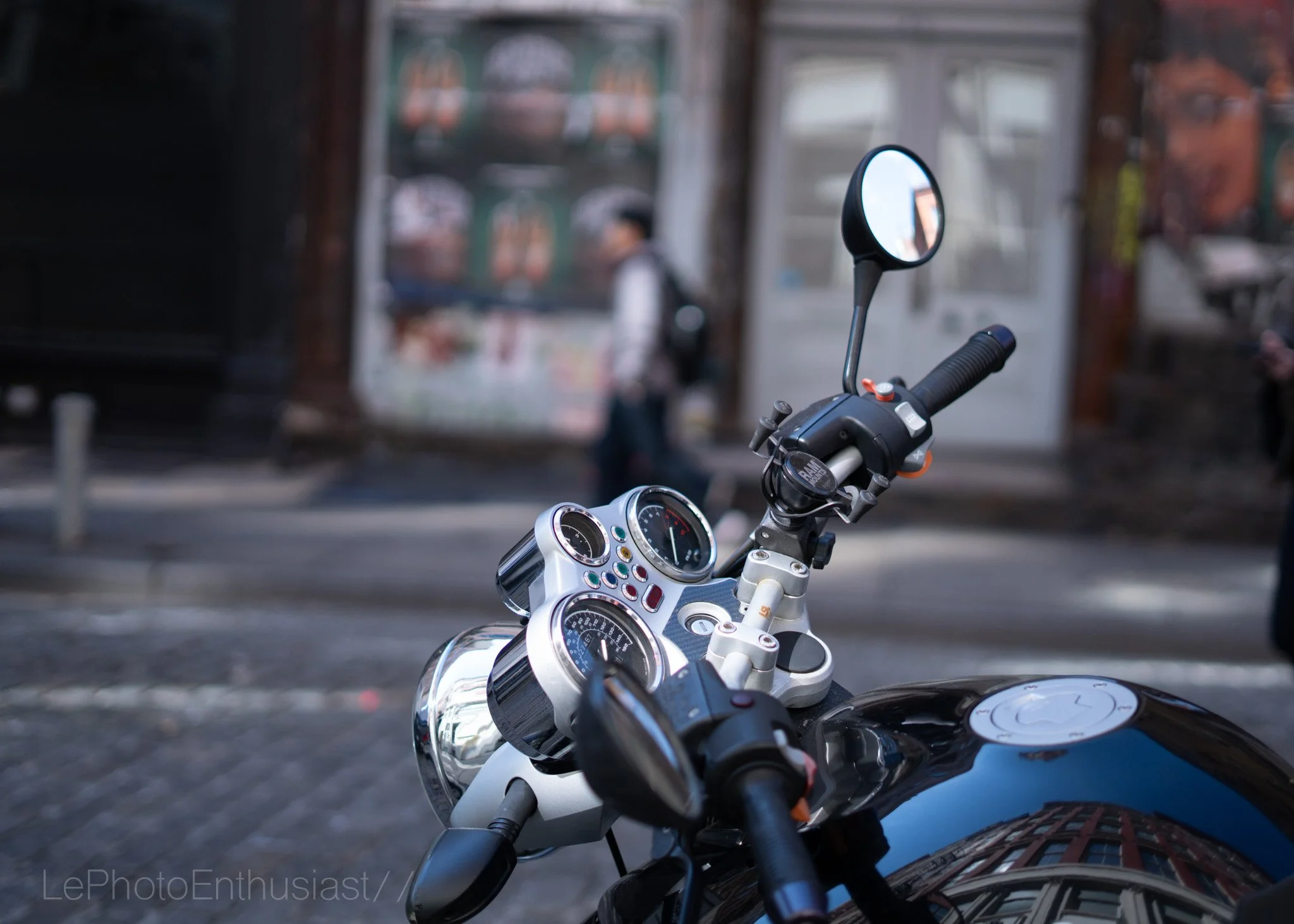 Close-up of motorcycle handlebars, rearview mirror, and dashboard in an urban setting with blurred buildings and a person walking in the background.