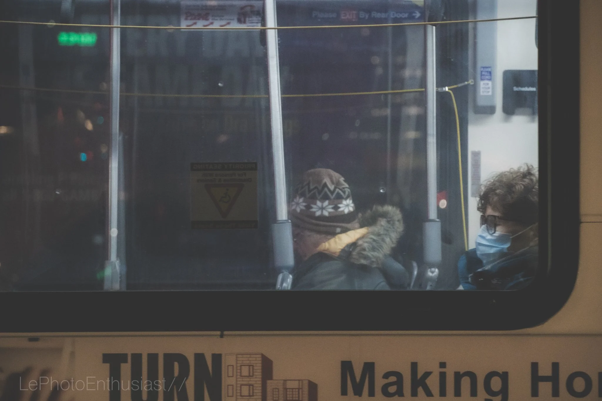 View through a bus window showing two passengers wearing masks, one with a winter hat and fur-lined hood, the other with glasses, in an indoor setting with signs and yellow strings overhead.