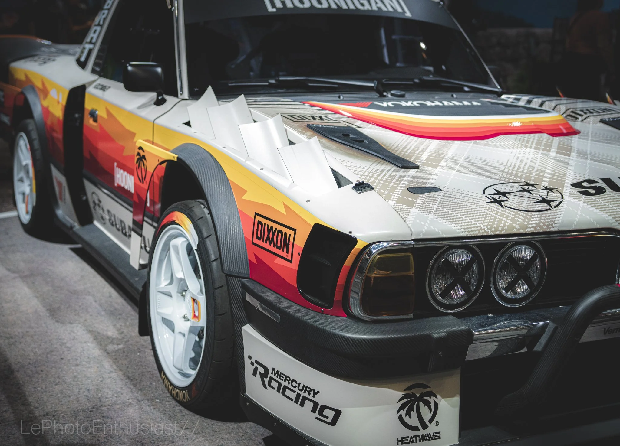 A race car with a colorful wrap featuring red, orange, and yellow flames, various sponsor logos including Mercury Racing, heatwave, and others, and a large windshield banner that reads 'KOSMIGAN'.