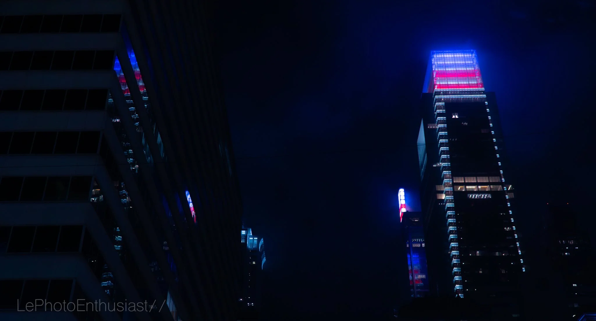Nighttime cityscape featuring a tall building illuminated with blue, pink, and purple lights.