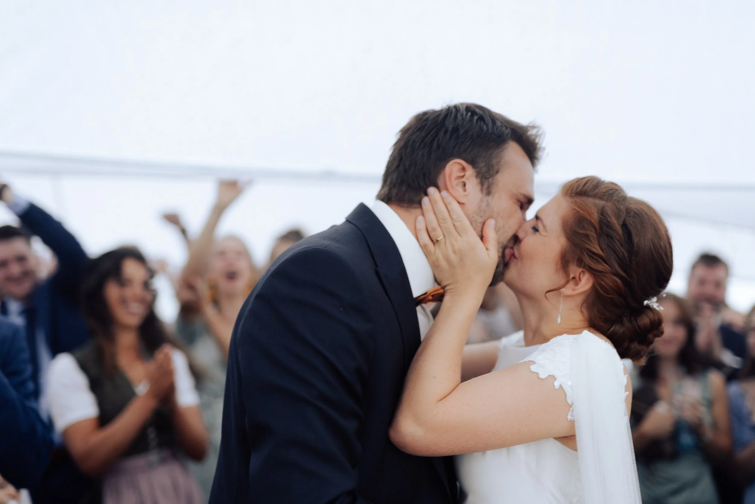 Ein Paar tanzt bei ihrer Hochzeit, sie küssen sich, im Hintergrund feiern Freunde.
