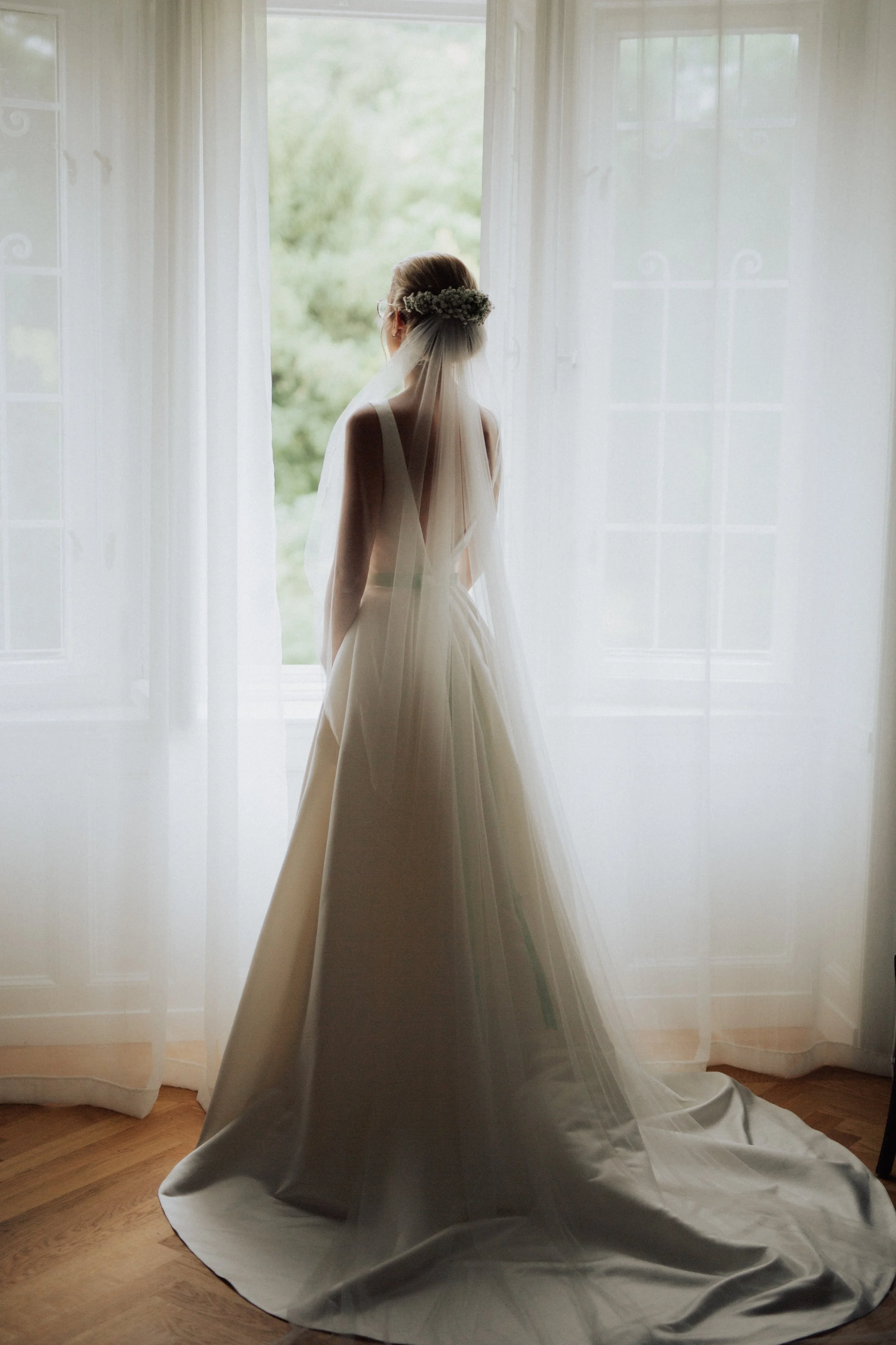 Braut in einem weißen Kleid mit Schleier und Blumenkranz blickt aus einem Fenster auf eine grüne Landschaft, im Hintergrund sind weiße Gardinen und Holzboden sichtbar.