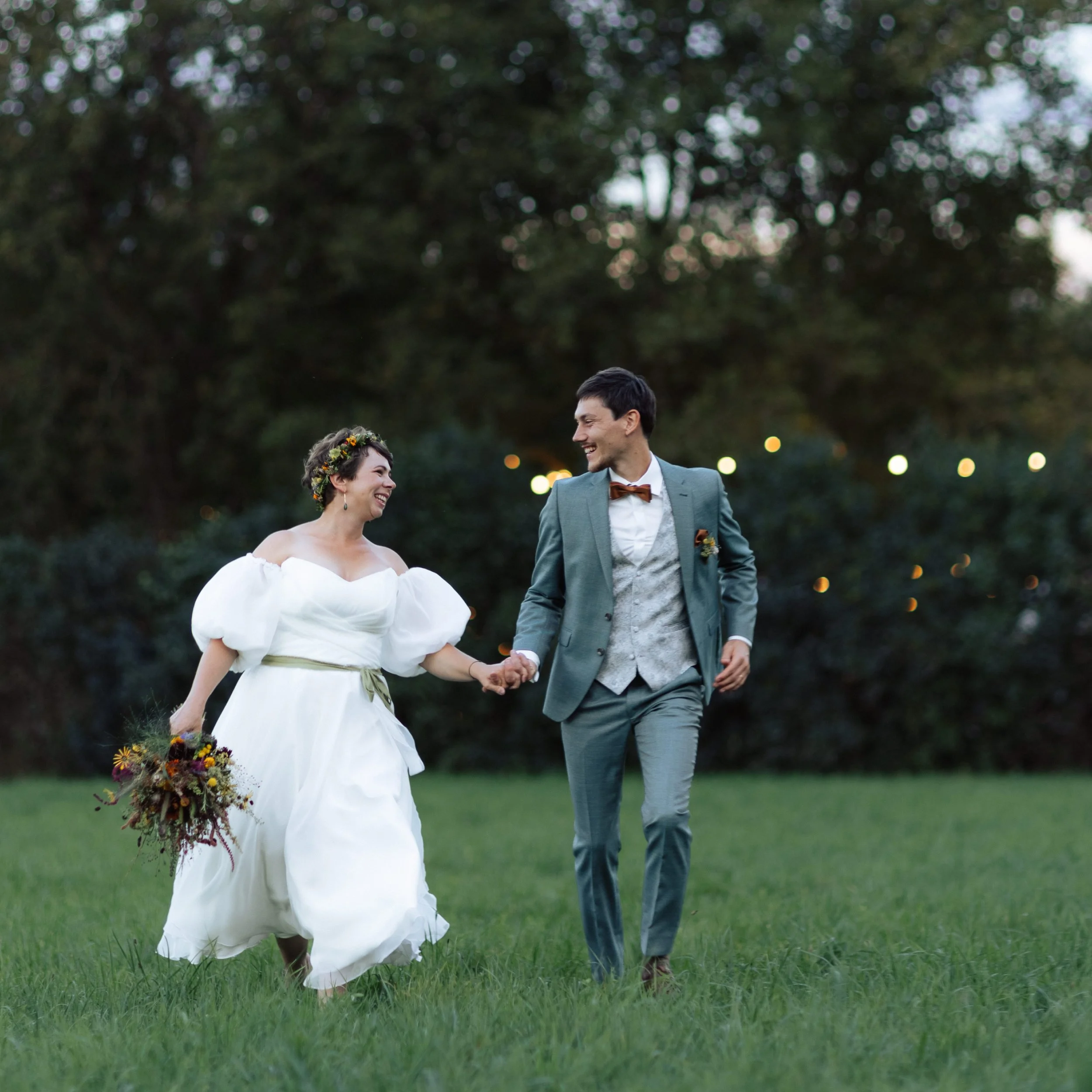 Ein lächelndes Paar bei einer Hochzeit im Freien, das miteinander tanzt, umgeben von grünen Bäumen und Lichterketten.