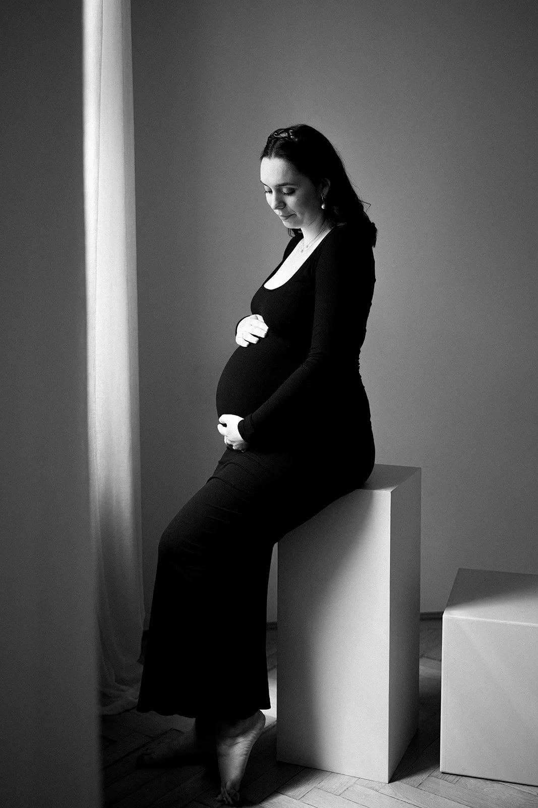 Schwangere Frau in schwarzem Kleid sitzt auf einem weißen Block, hält den Babybauch, blickt nach unten, schwarz-weiß.