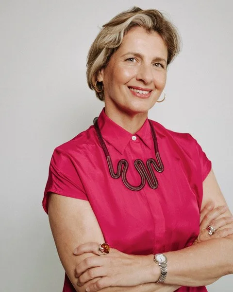 A woman with short light brown hair styled in a bob, wearing a colorful blouse with an abstract pattern of circles in red, black, white, beige, and gold, and a matching tie at the neckline, smiling against a plain grey background.
