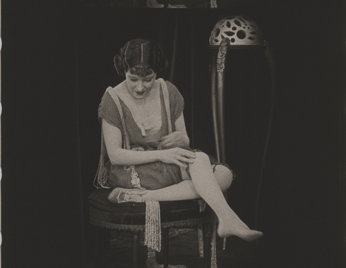 Black and white photo of a woman seated on a stool wearing a vintage dress, adjusting her legwear. An ornate stand is in the background.