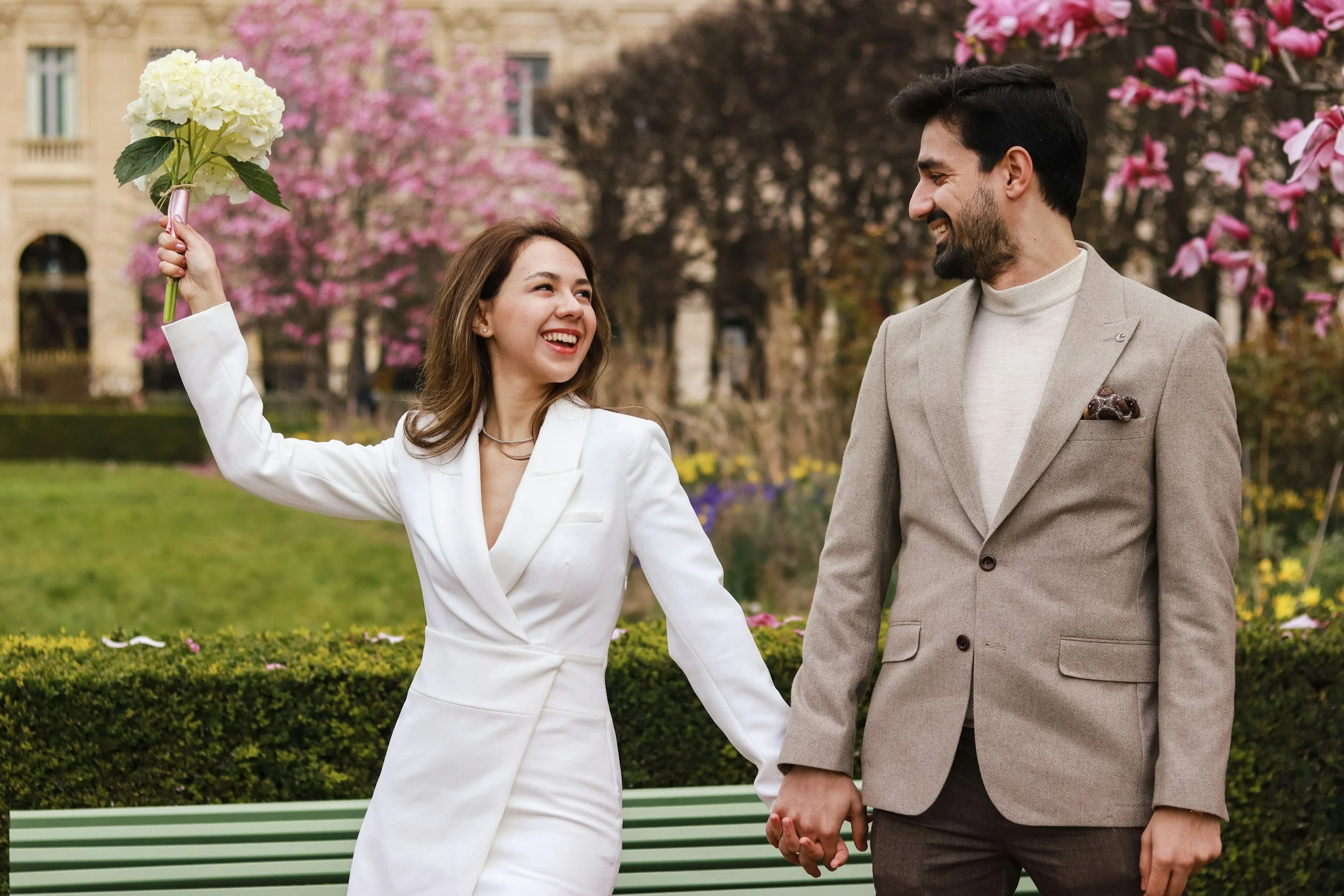 A woman and man holding hands in a park with pink flowering trees. The woman is wearing a white suit and holding a bouquet of white flowers, smiling at the man, who is dressed in a beige suit with a pocket square, smiling back.