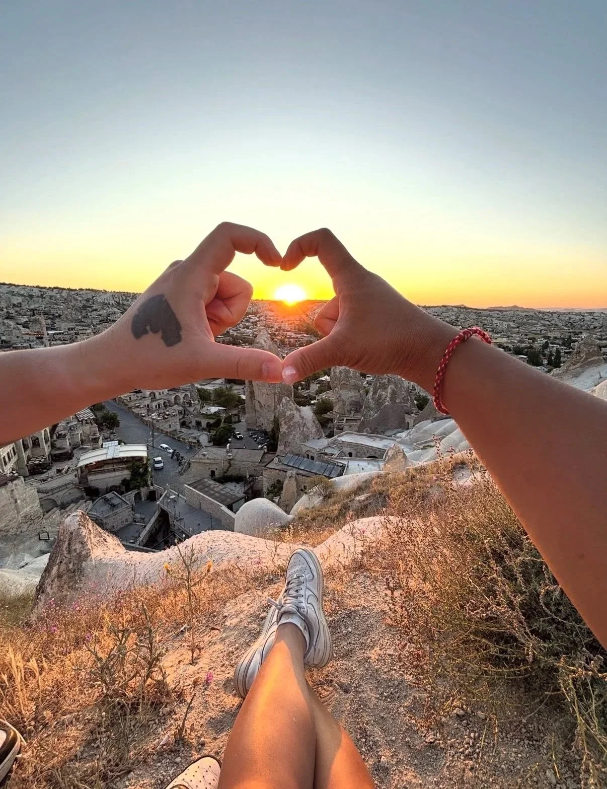 Two hands forming a heart shape around a setting sun over a cityscape. One hand has a tattoo, and the other is wearing a red bracelet. The person's legs and shoes are visible in the foreground, sitting on a rocky hillside with dry grass and small plants.
