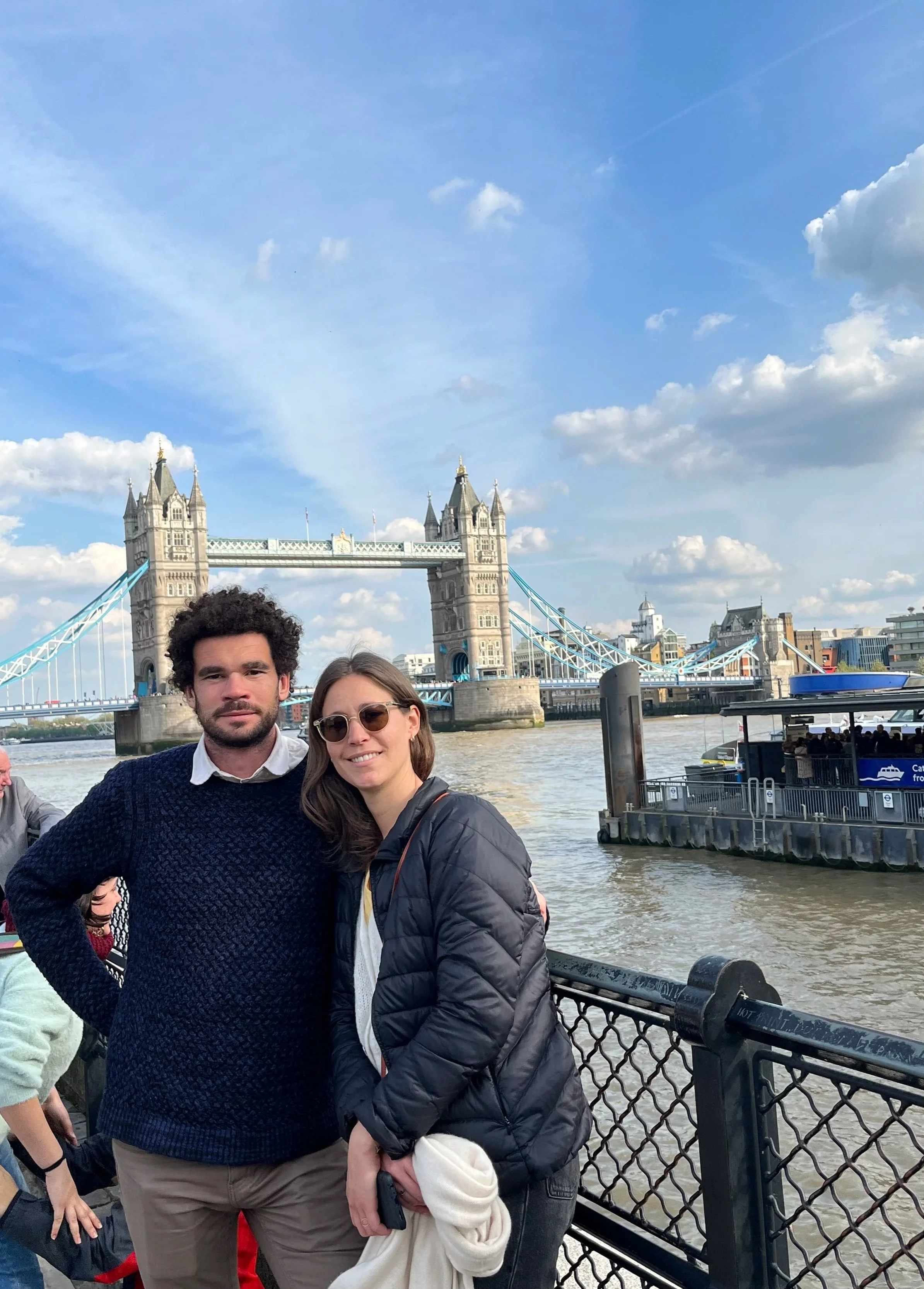 Two people are standing close to each other on a bridge by the river in London with the Tower Bridge in the background, under a partly cloudy sky.