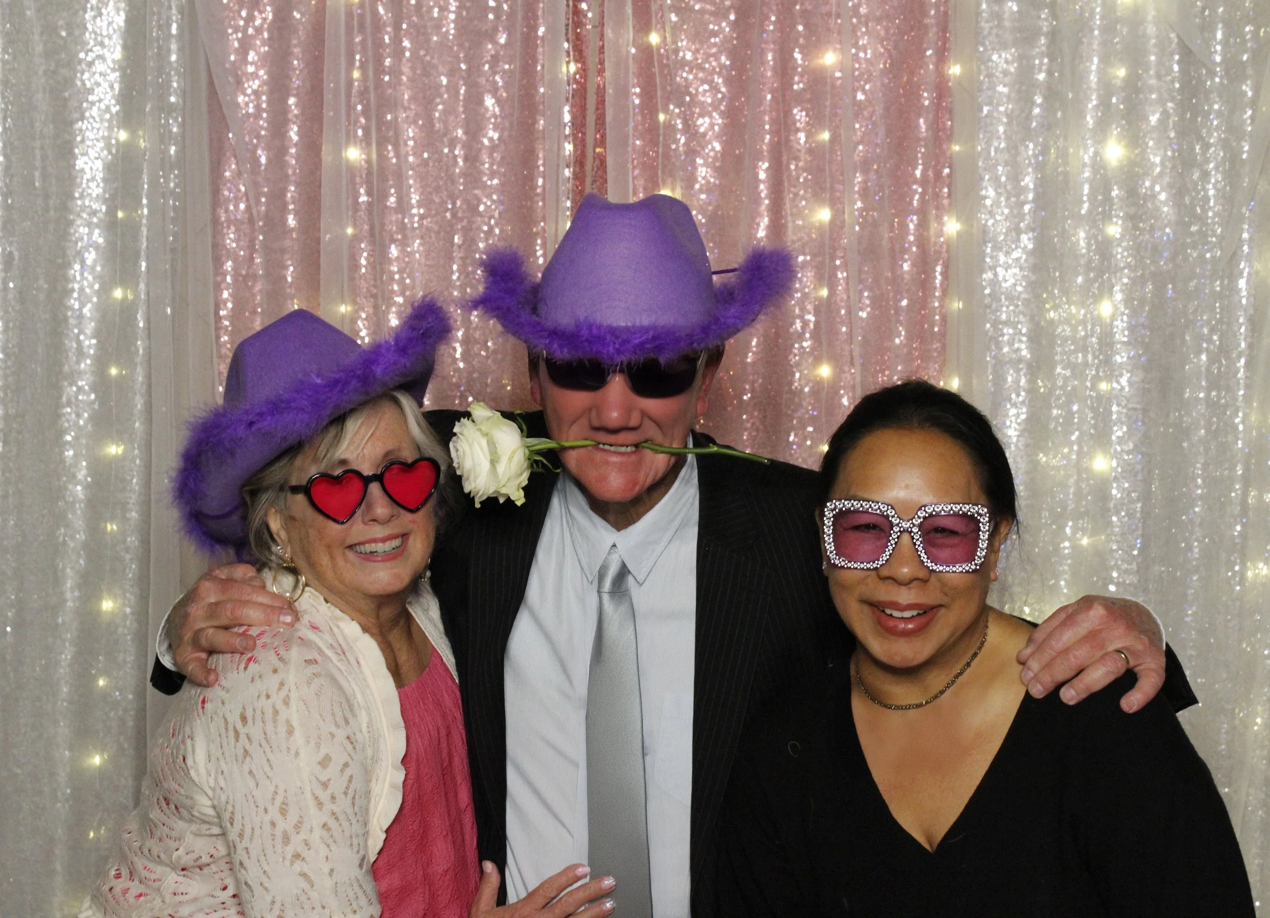 Four people posing together at a festive event with sparkly backdrop. They are wearing fun accessories including large glasses and purple hats, and one person has a white rose in mouth. Smile and embrace each other.