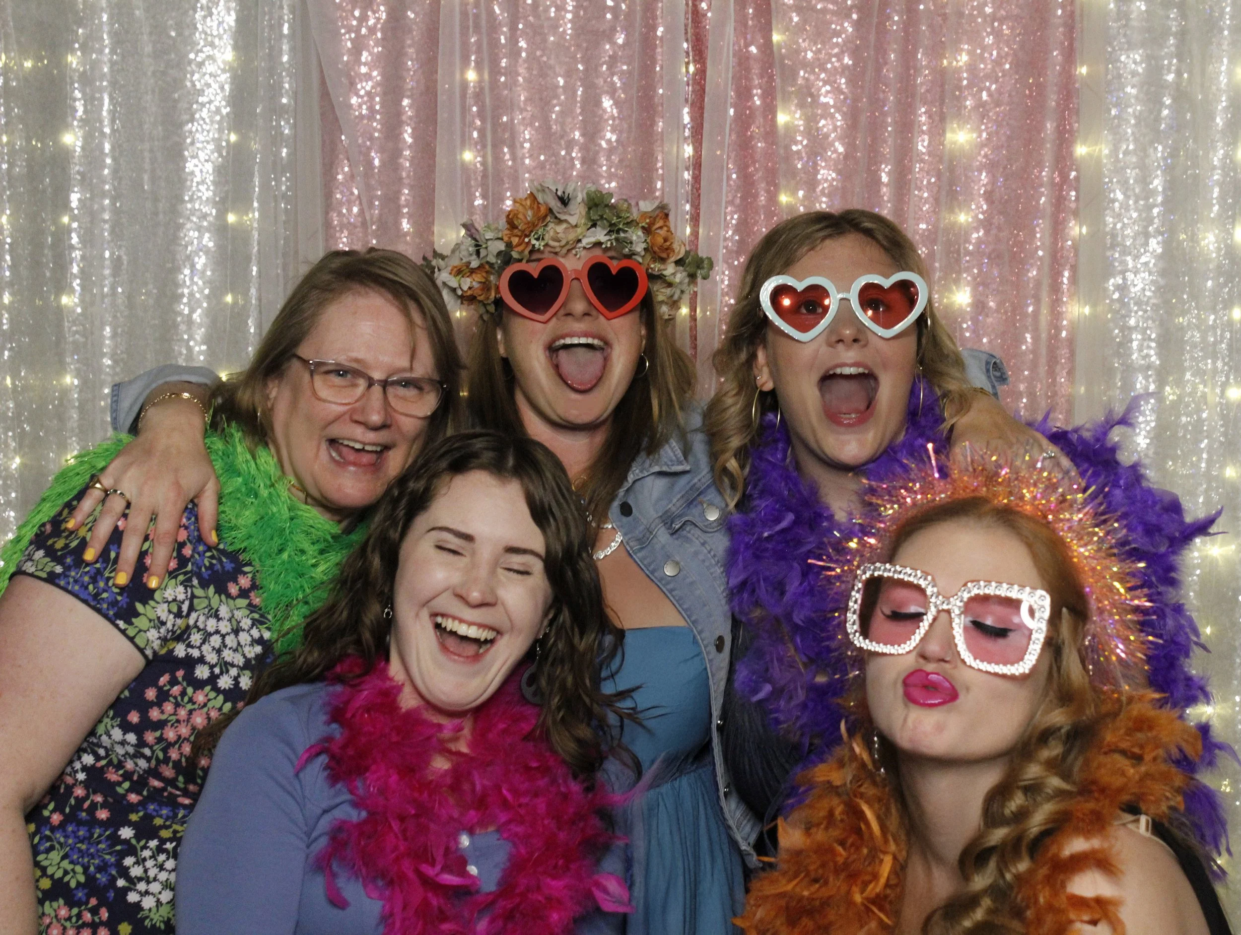 A group of women at a celebration or party, wearing colorful feather boas, large sunglasses, and accessories, with a sparkly pink and white curtain backdrop.