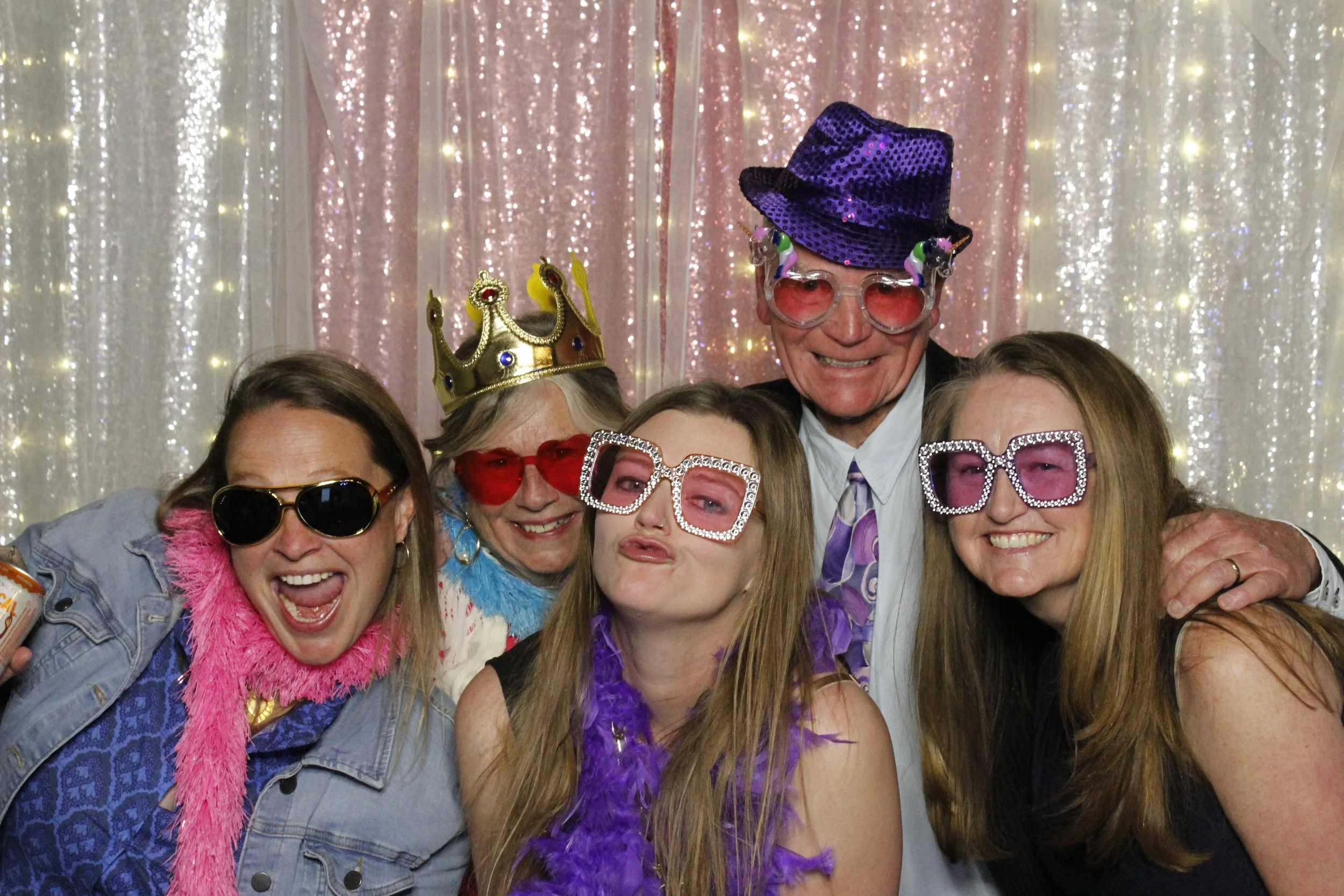 Group of people celebrating, wearing colorful costumes, glasses, hats, and feather boas, with a sparkling curtain background.