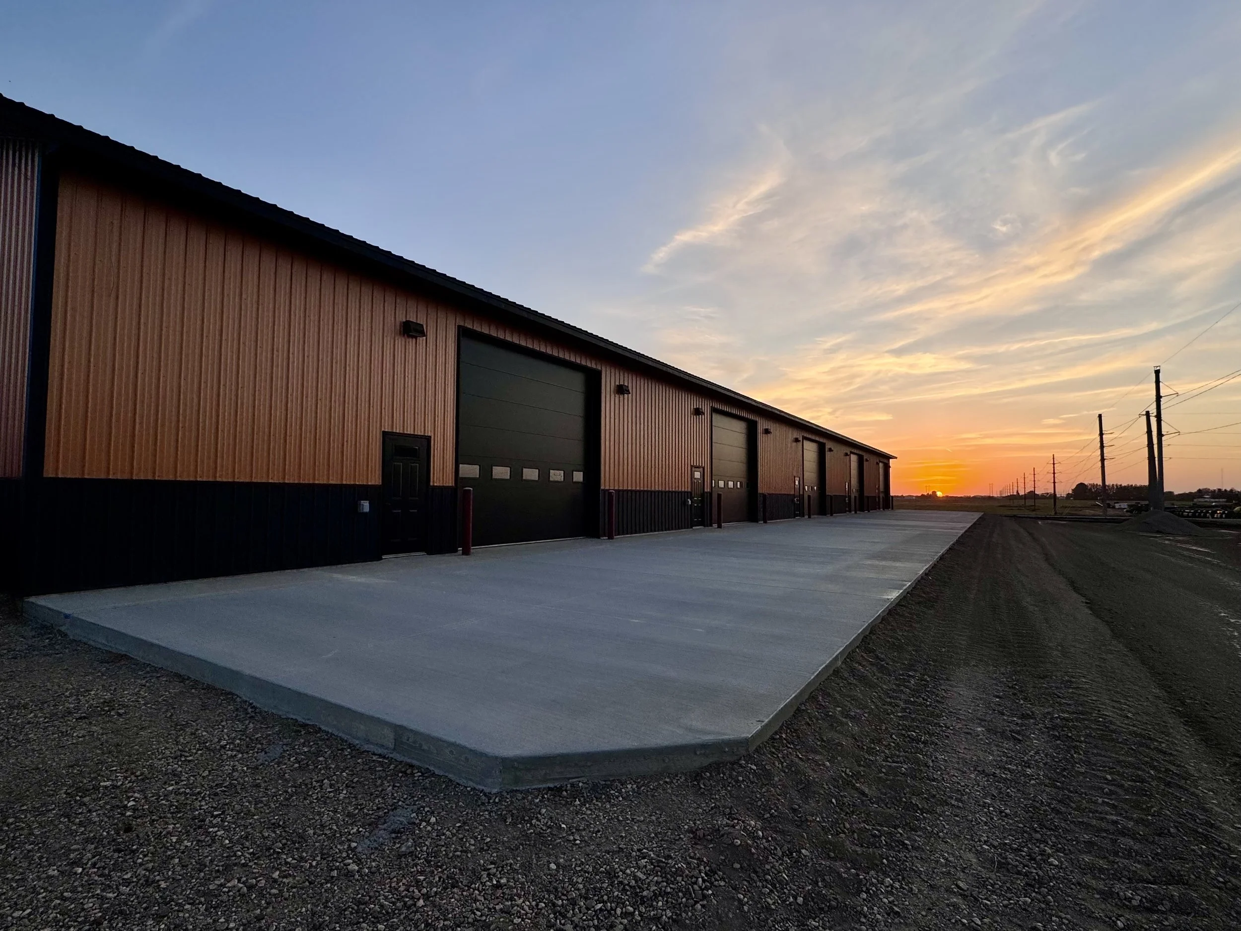 A row of large black garage doors on a building with wood siding, set against a sunset with orange and blue sky, and power lines in the background.