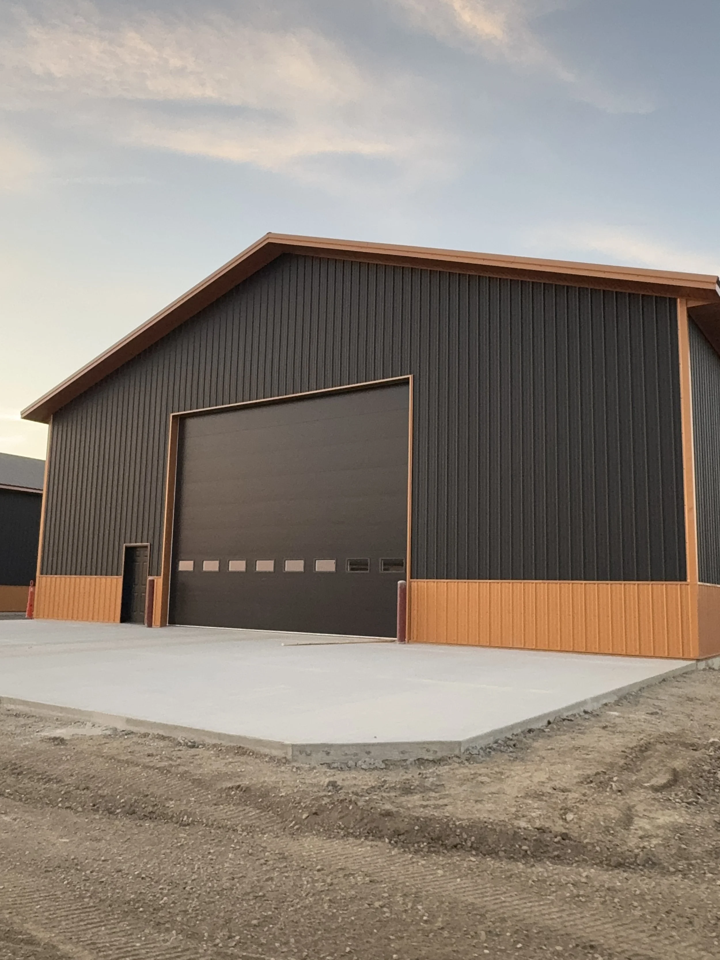 A modern, black and wood-paneled industrial building with a large garage door and a smaller door, situated on a concrete slab with dirt around it, during dusk or dawn.