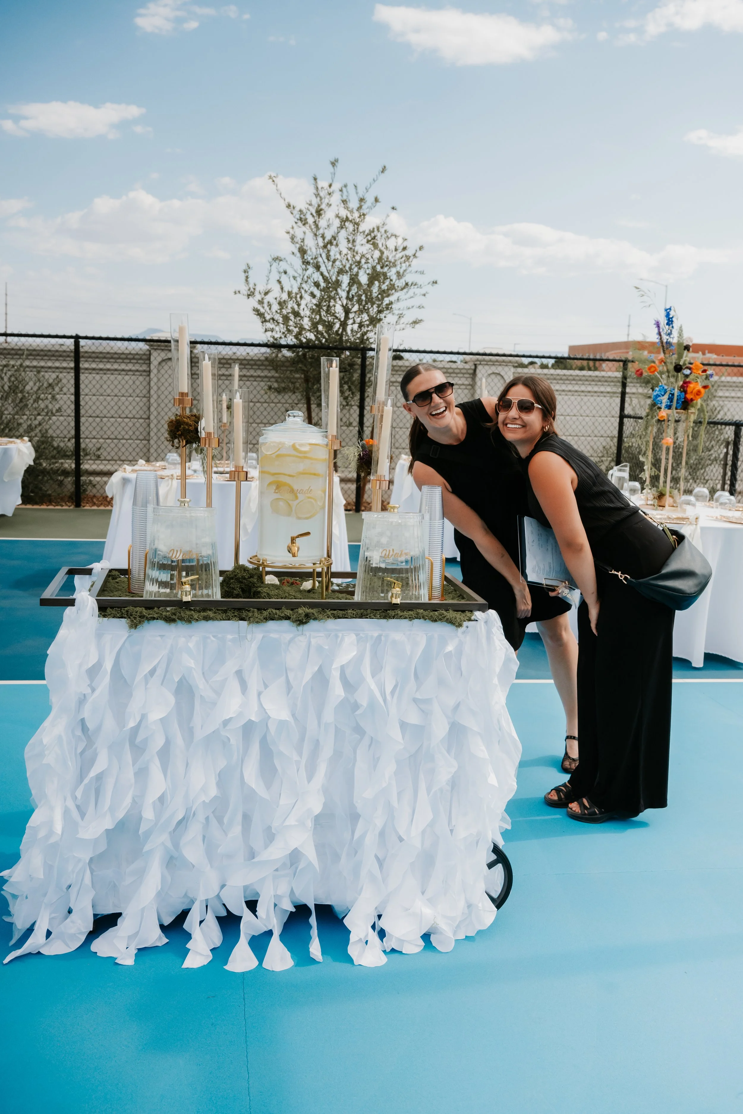 Two women in black dresses smiling and leaning towards each other at an outdoor event, with a table featuring a large drink dispenser, glasses, candles, and a flower arrangement in the background.