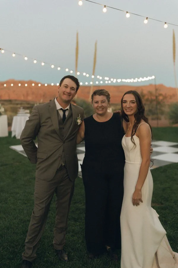 People at an outdoor evening event, likely a wedding reception, standing on grass under string lights with a scenic desert landscape in the background.