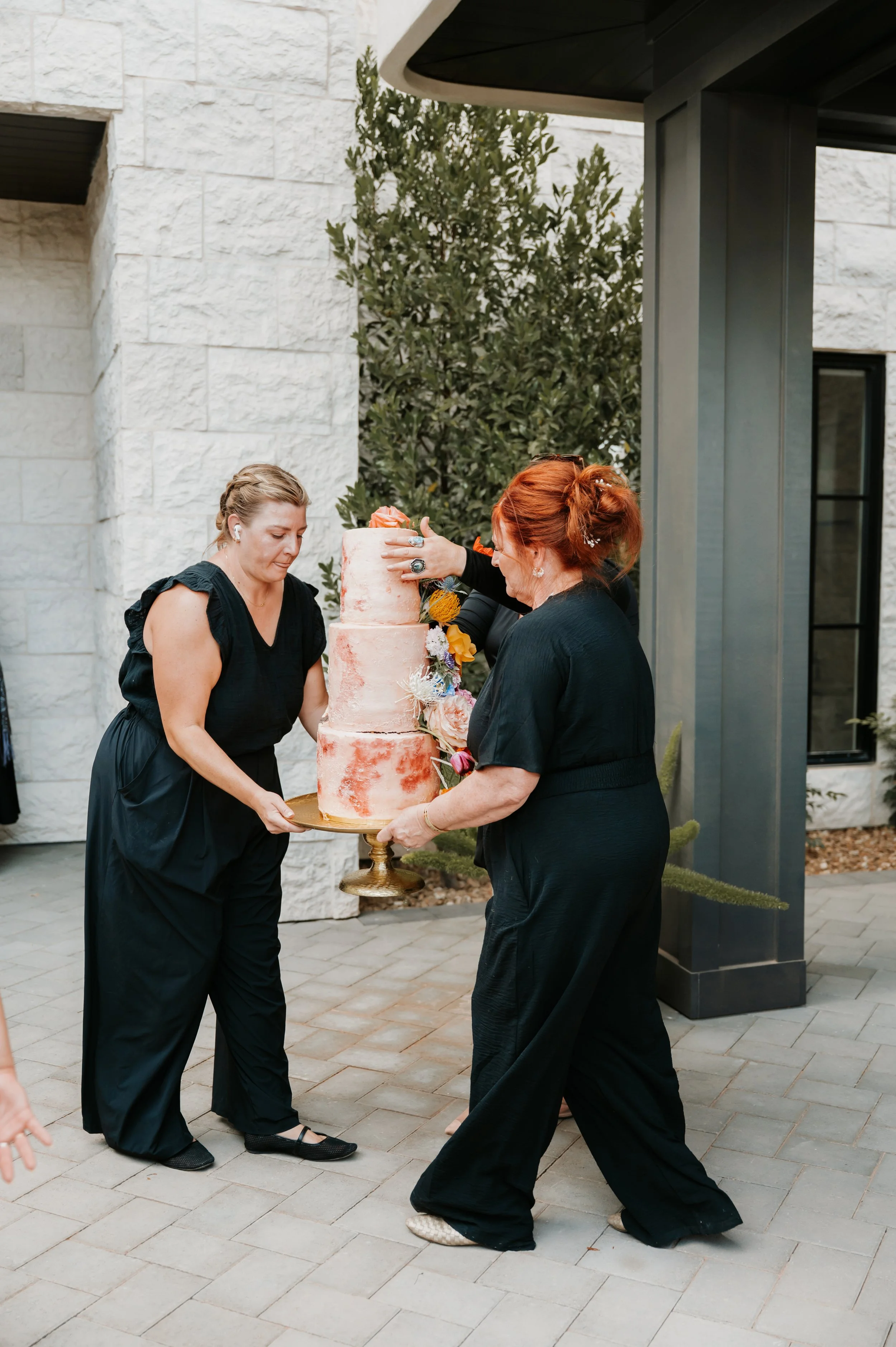 Two women dressed in black are preparing to cut a tall, three-tiered pink and orange ombre wedding cake decorated with flowers. They are outdoors on a paved patio near a white stone building with greenery in the background.
