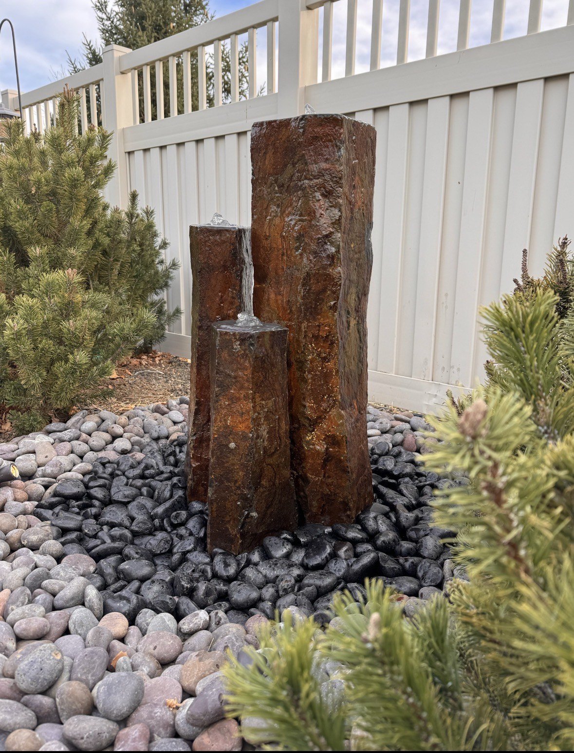 Three vertical rust-colored stone water fountains of varying heights with water flowing from the top, placed on black pebbles, surrounded by greenery, in front of a white fence.