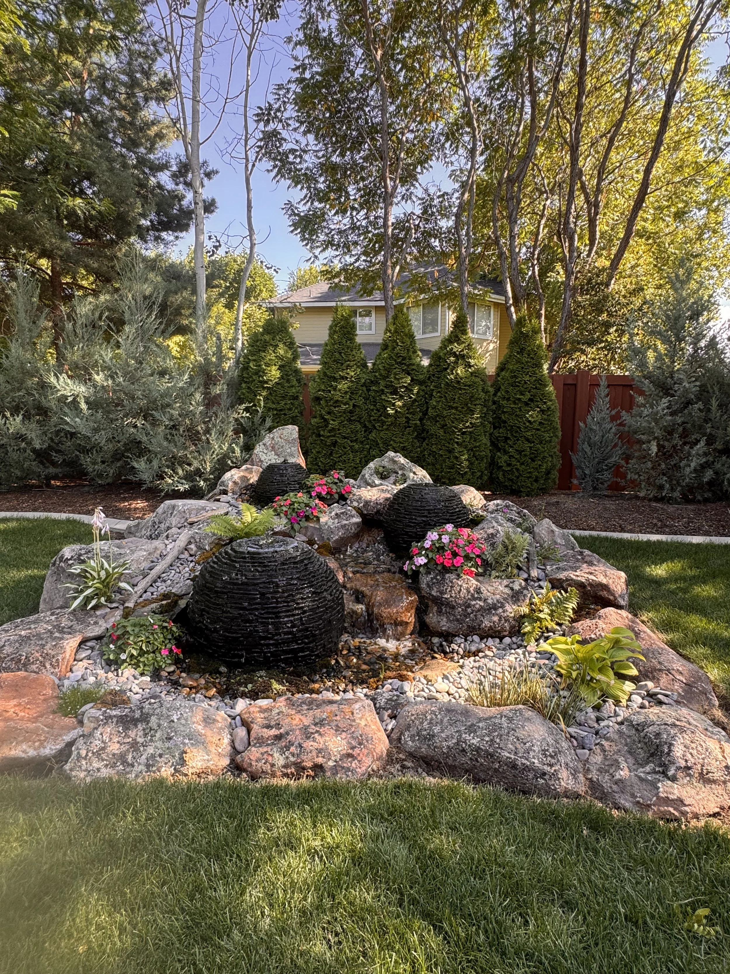 A landscaped backyard garden with a rock fountain, pink and purple flowers, green shrubs, and a red wooden fence in the background, with a house and trees overhead.