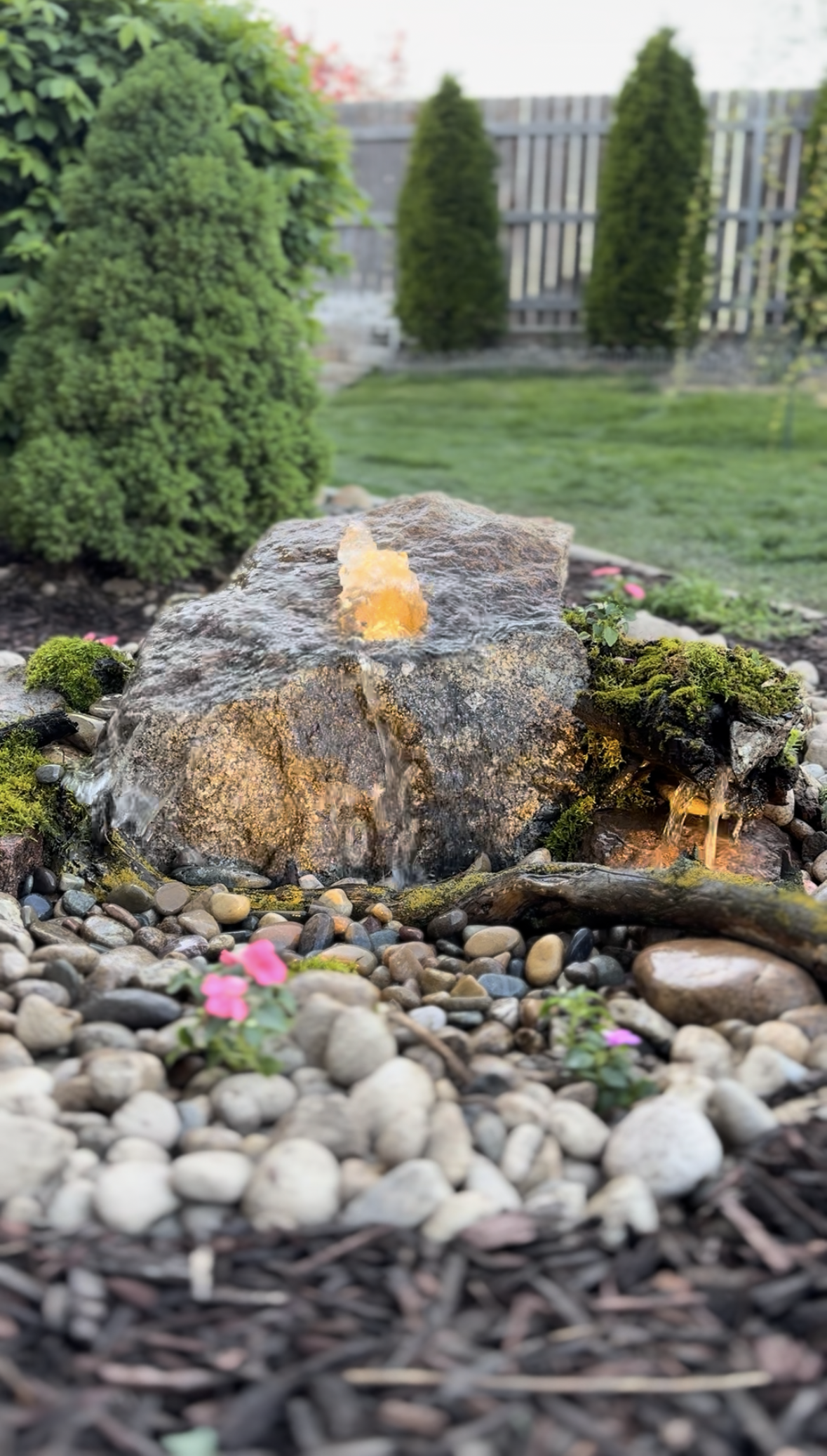 A landscaped garden with a small water fountain built around a large rock, with moss and flowers, surrounded by pebbles and greenery.