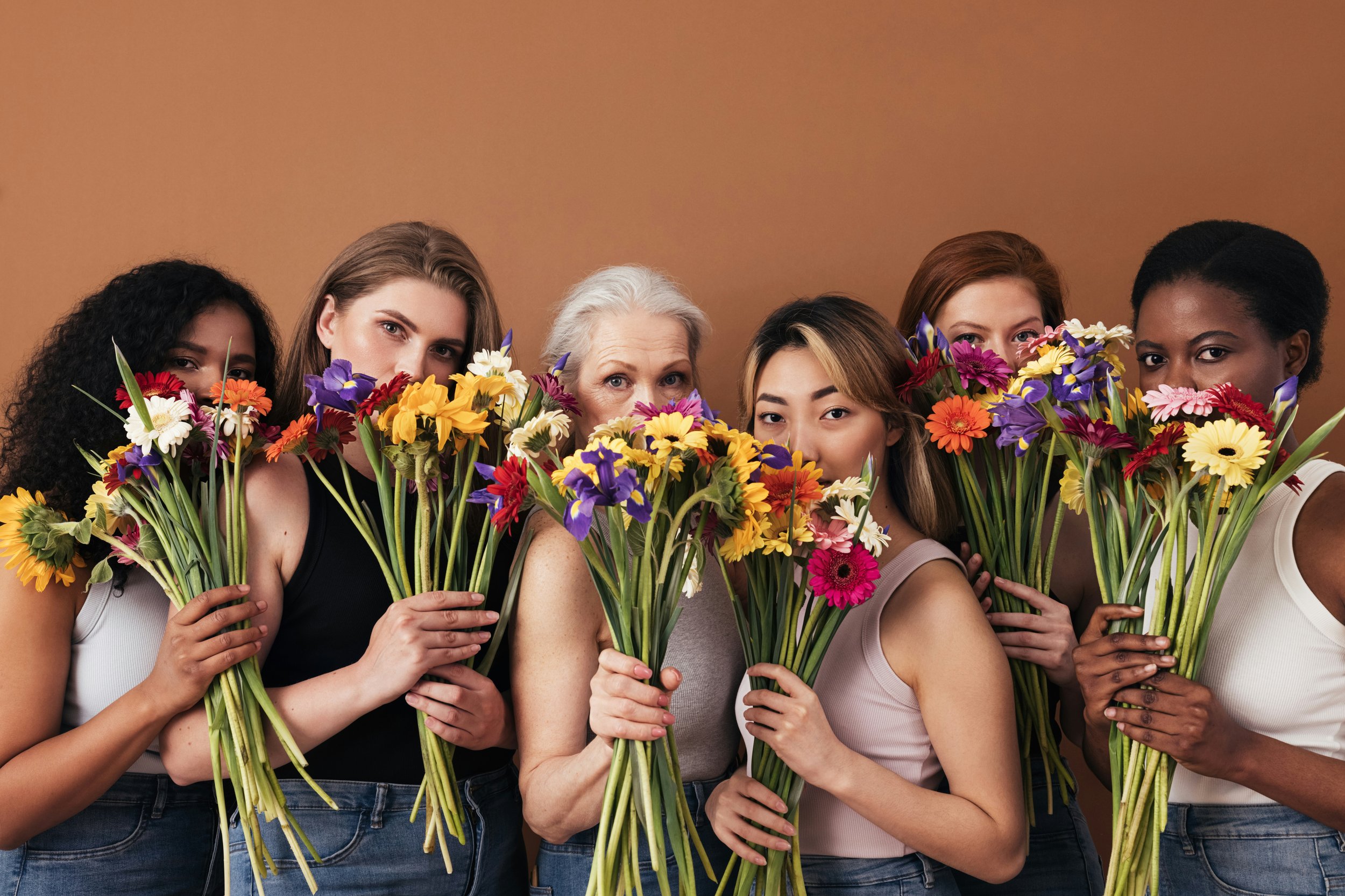 Six women holding colorful bouquets of flowers, partially obscuring their faces, standing against a plain brown background.