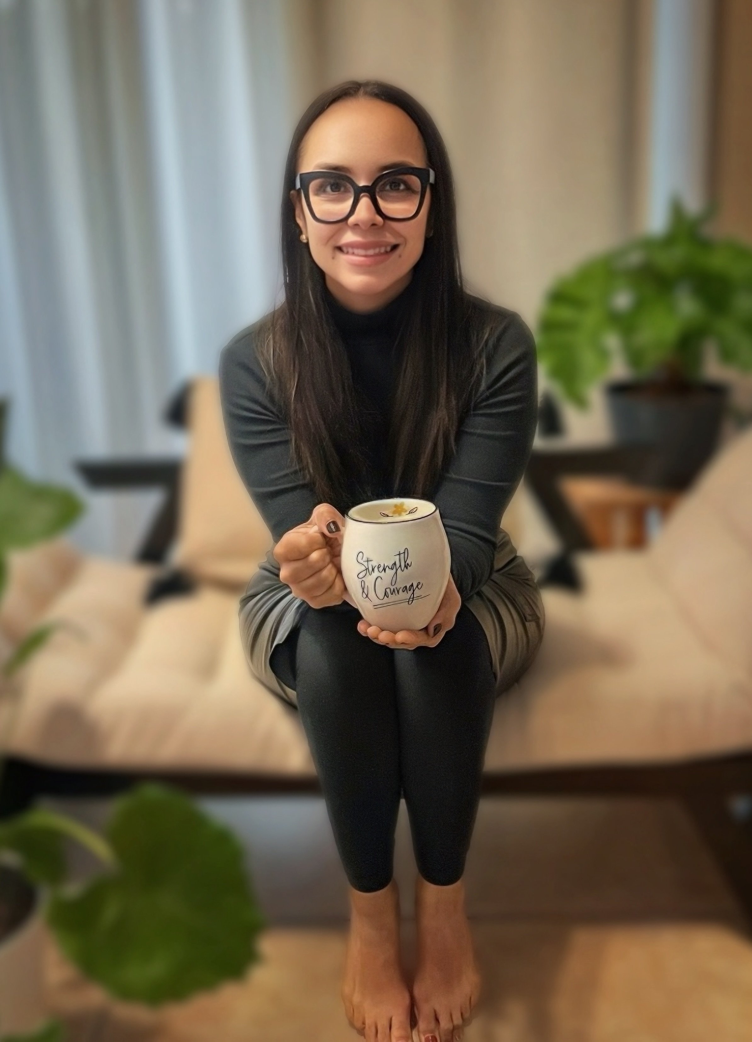 A woman with long dark hair, glasses, wearing a black top and beige skirt, sitting on a beige couch holding a white mug with the words "Strength & Courage" written on it. There are green plants in the background.