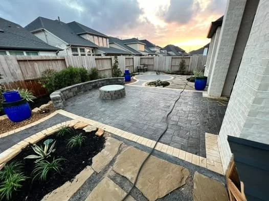 View of a backyard patio with newly installed pavers, surrounded by garden beds with green plants, large blue planters, and a wooden fence, during sunset.